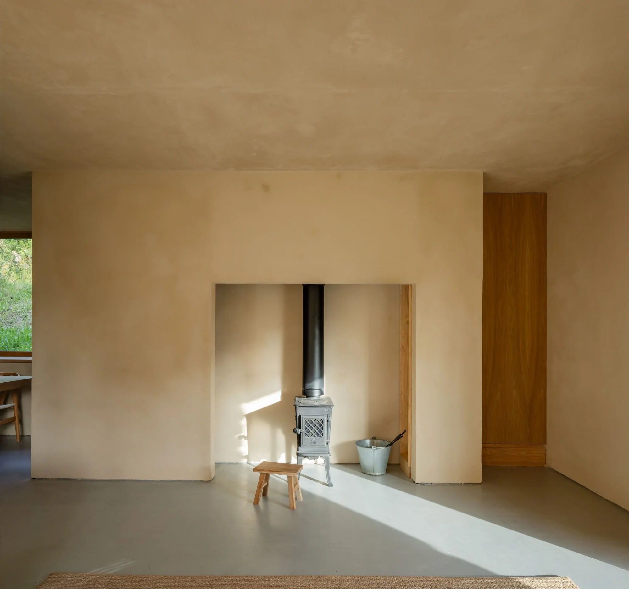 View of a minimalist fireplace with light wood cabinets, decorative objects and plants on a shelf, and a wooden table with wooden chairs next to a beige wall.