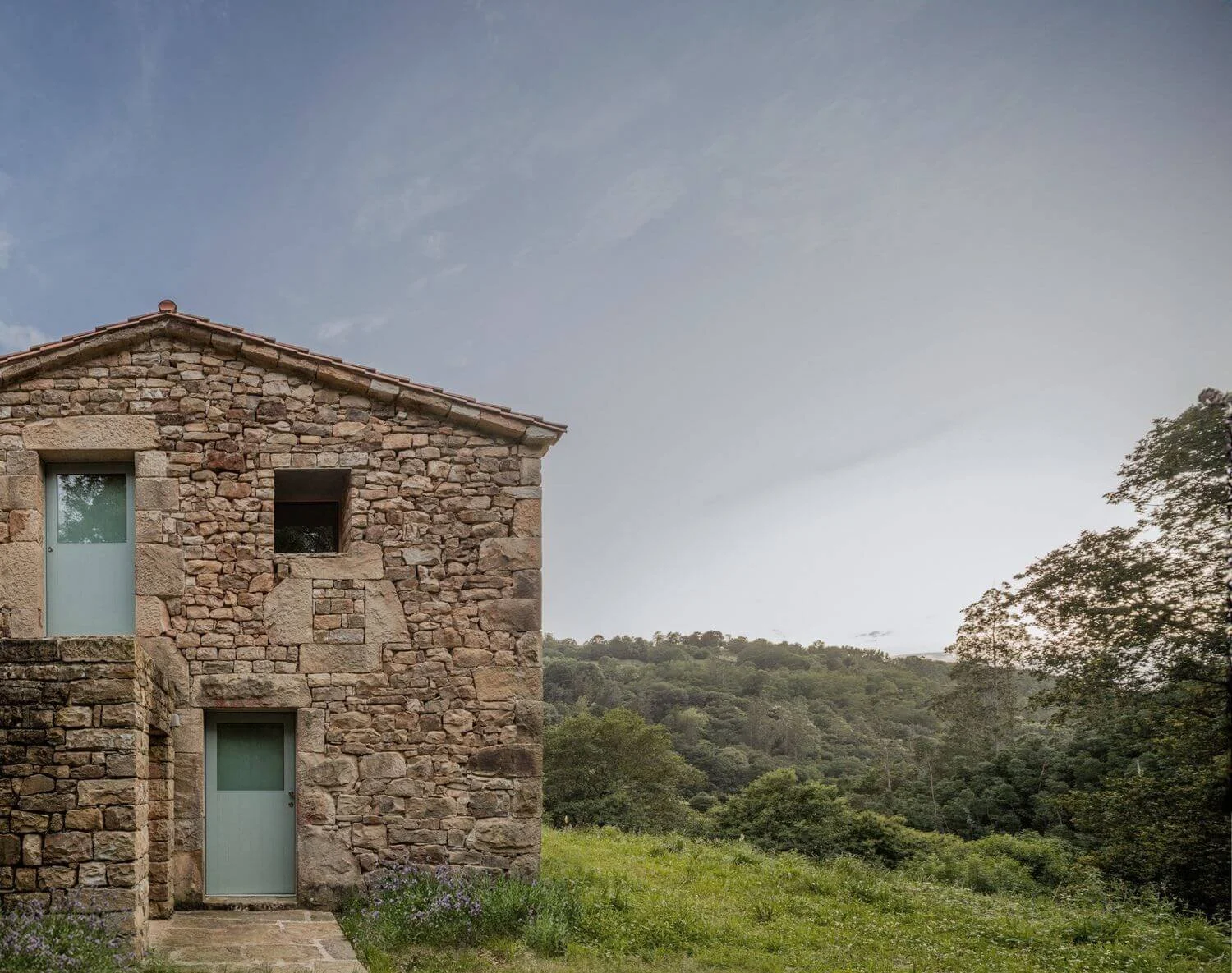 Casa Passivhaus de piedra en un campo con árboles y camino, cielo con nubes.