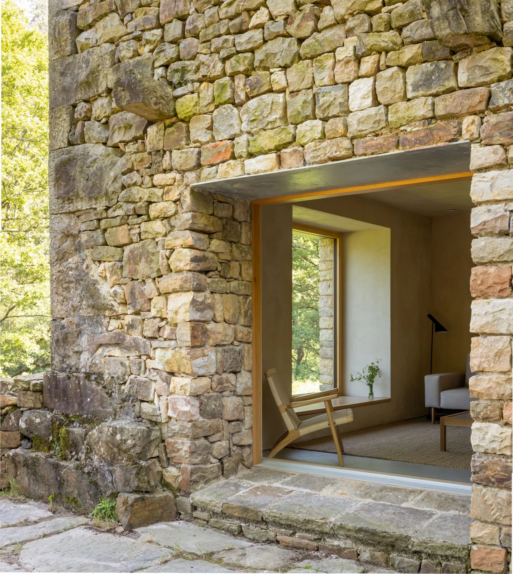 Vista del interior de una casa con paredes de piedra, un sillón de madera y tela, una lámpara de pie negra, y una ventana que muestra árboles verdes afuera.