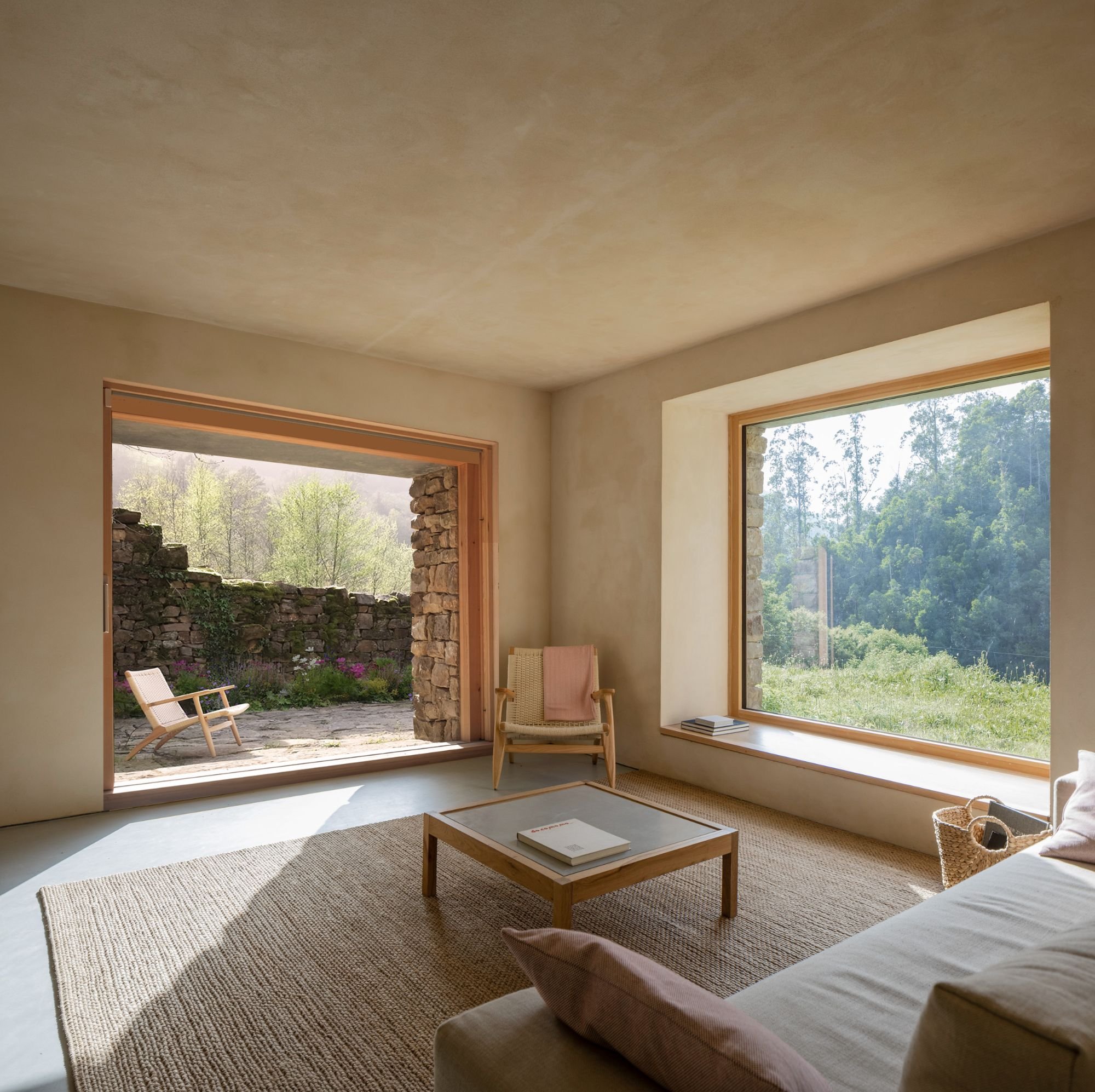 Interior of a living room with large windows showing a landscape of green trees and mountains, a wooden chair with a cushion, a black floor lamp, and a vase of plants on a wooden table.