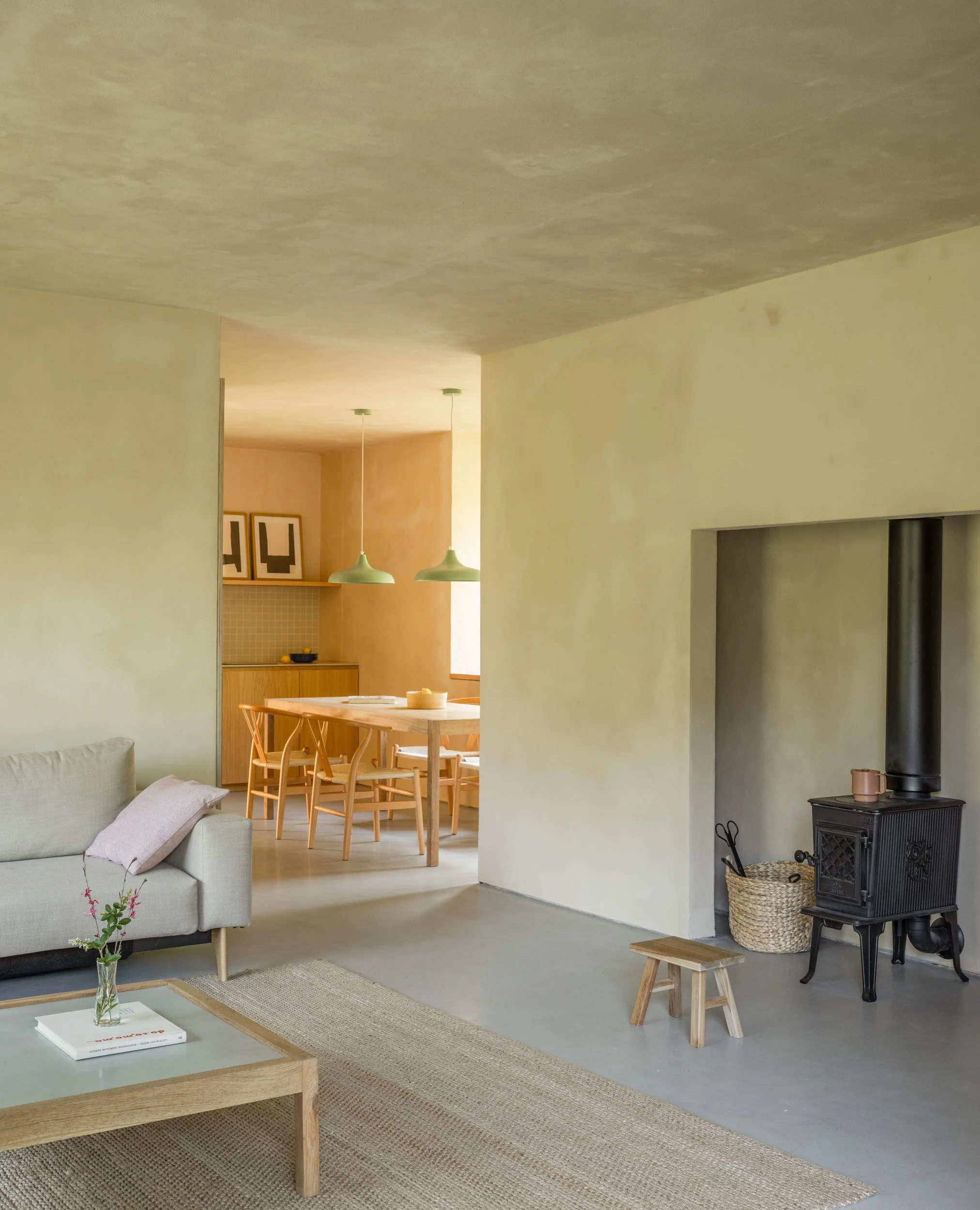 Vista de una cocina minimalista con gabinetes de madera clara, objetos decorativos y plantas sobre una repisa, y una mesa de madera con sillas de madera junto a una pared beige.