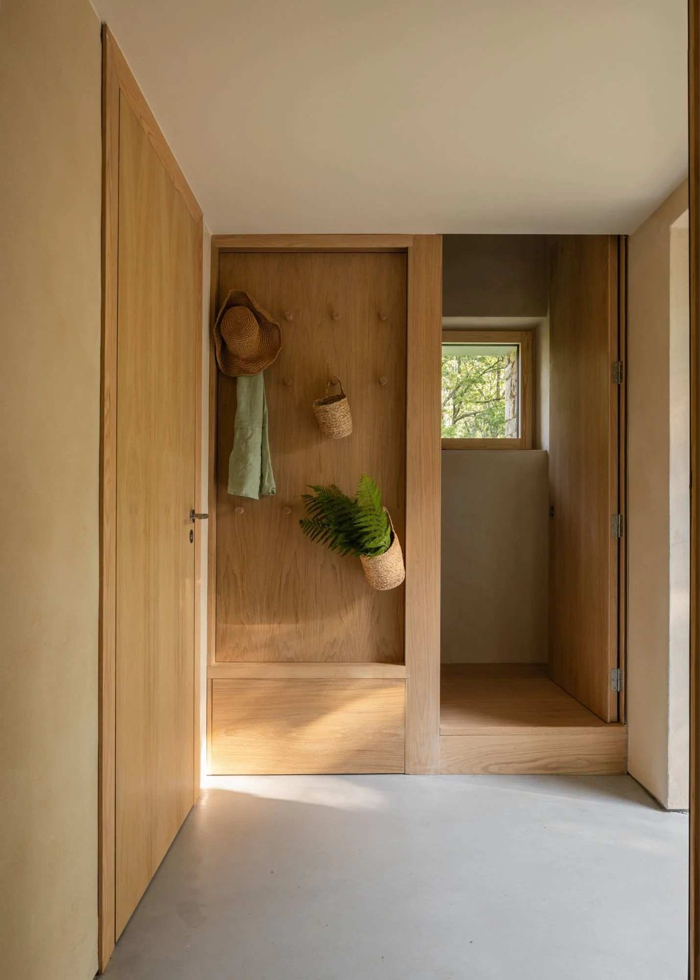 Entrada con paredes y puertas de madera, perchero con sombrero, tela y plantas en cestas, ventana que muestra árboles afuera.