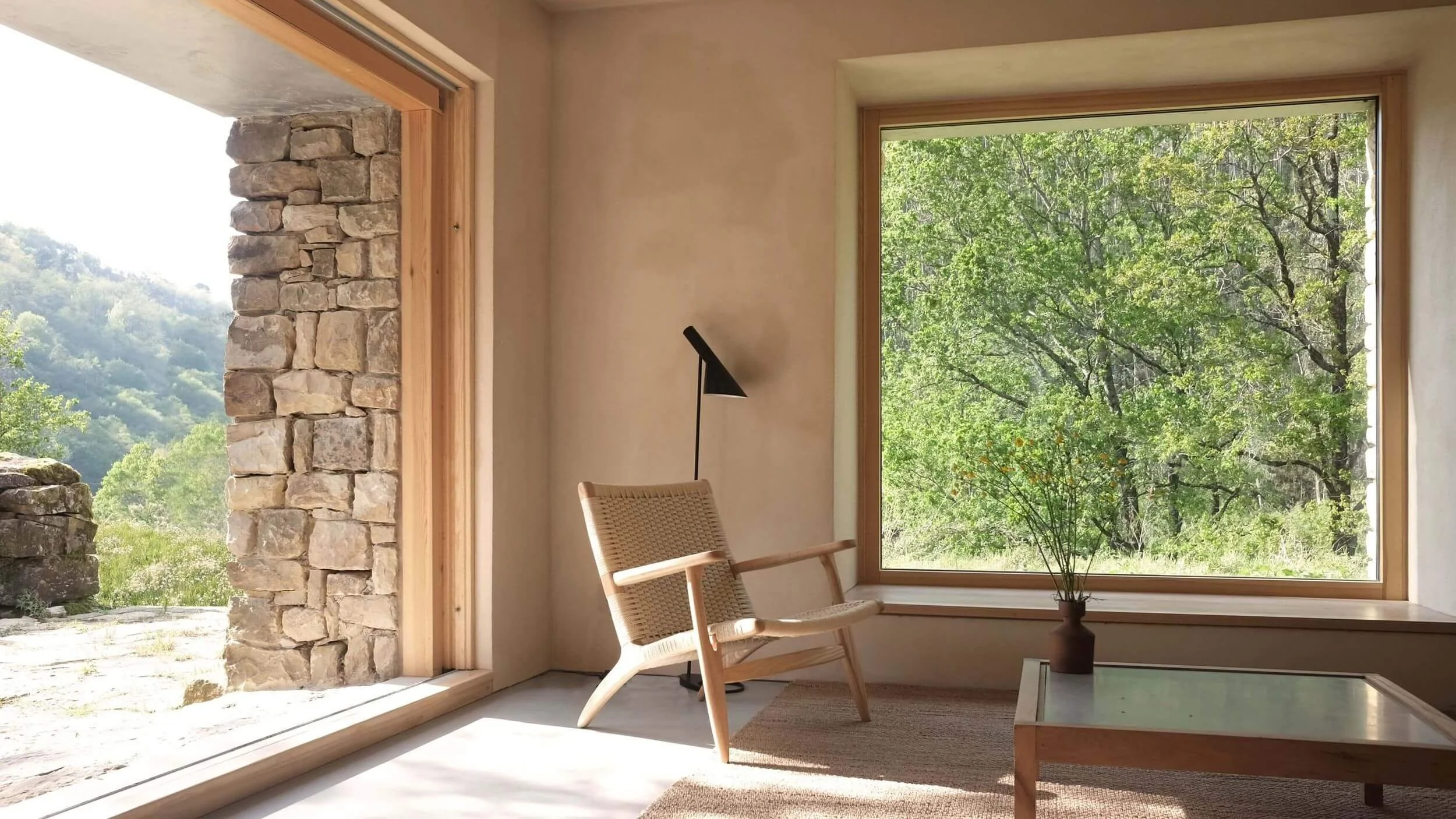 Interior de una sala con gran ventana mostrando árboles verdes y paisaje montañoso, sillón de madera y mimbre, lámpara de pie negra, mesa de centro de madera con jarrón y plantas, luz natural y ambiente minimalista.