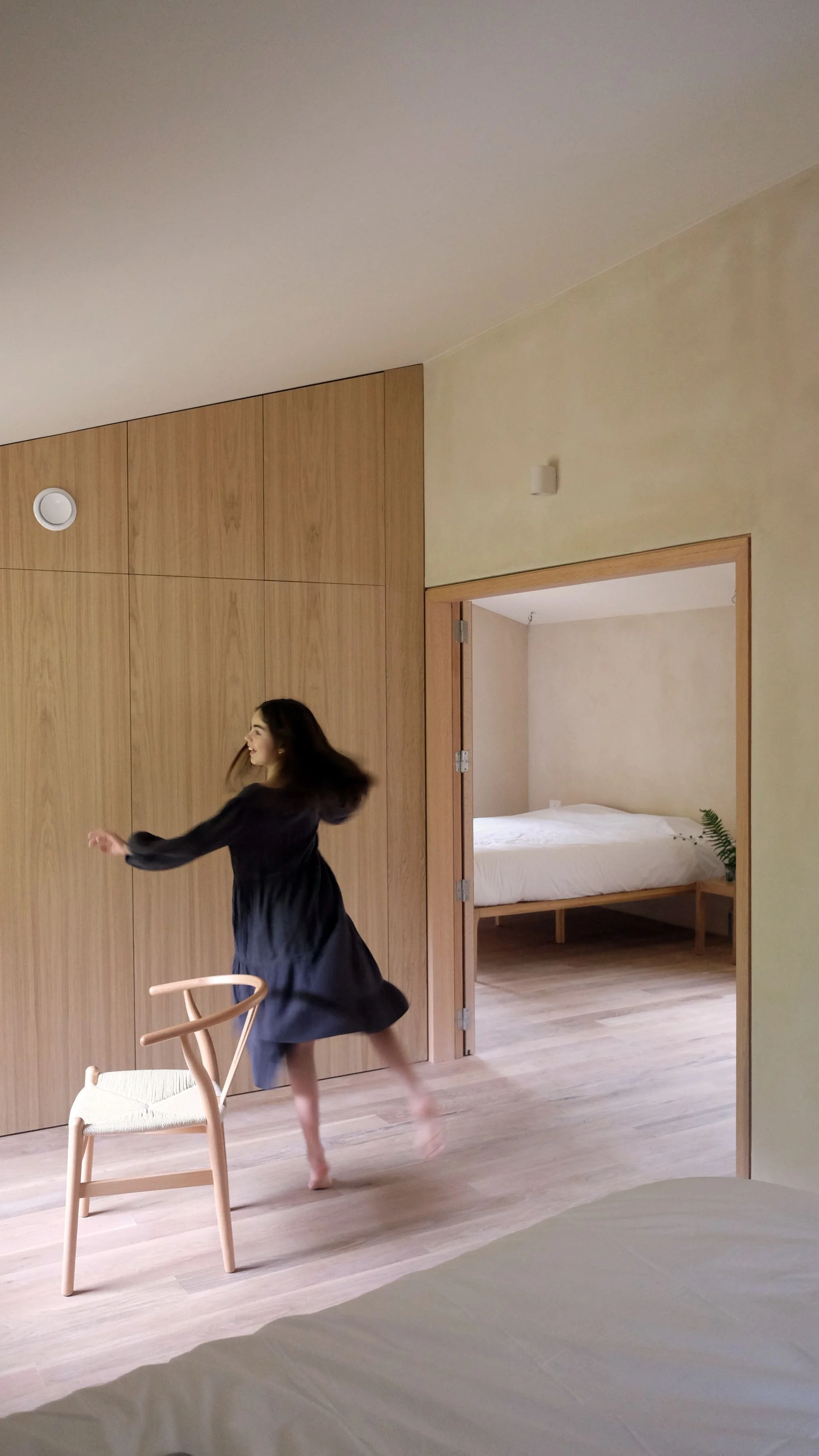 A girl in a black dress appears to be dancing or spinning in a modern room with wooden walls and light wood floors, near a chair and an additional space with a bed and a plant.