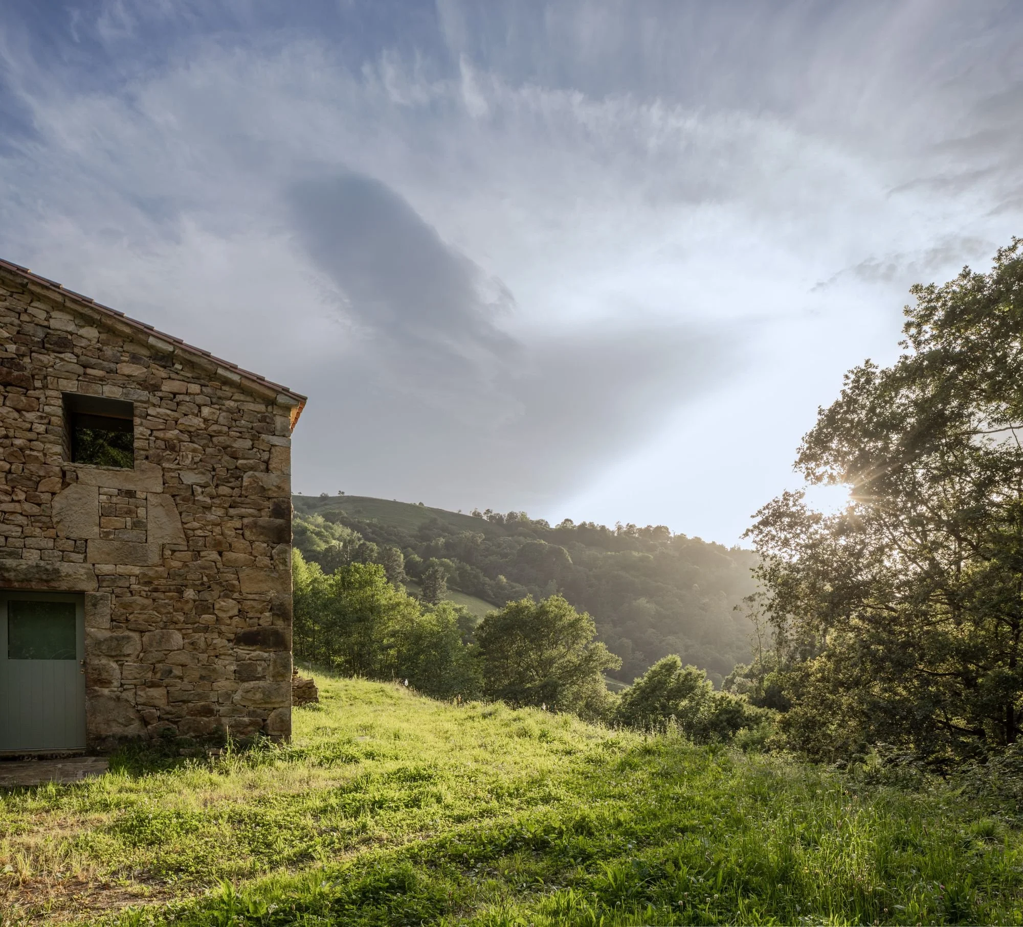 Vista de un paisaje con una casa de piedra, árboles verdes y colinas al fondo durante el atardecer.