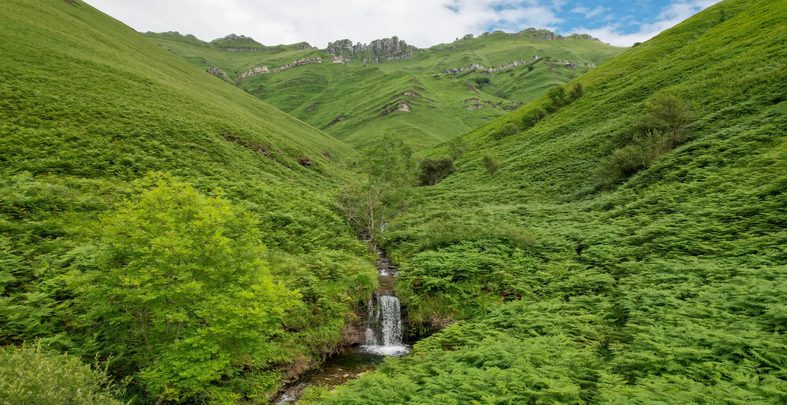 Paisaje natural con colinas verdes cubiertas de vegetación, un pequeño río con cascada en el centro y montañas en el fondo, cielo parcialmente nublado.
