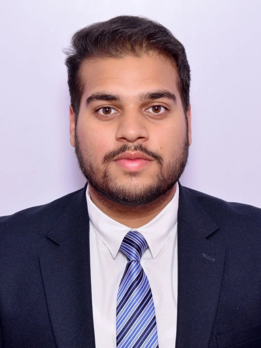 A man with dark hair and a beard wearing a black suit, white shirt, and striped tie, looking directly at the camera against a plain white background.