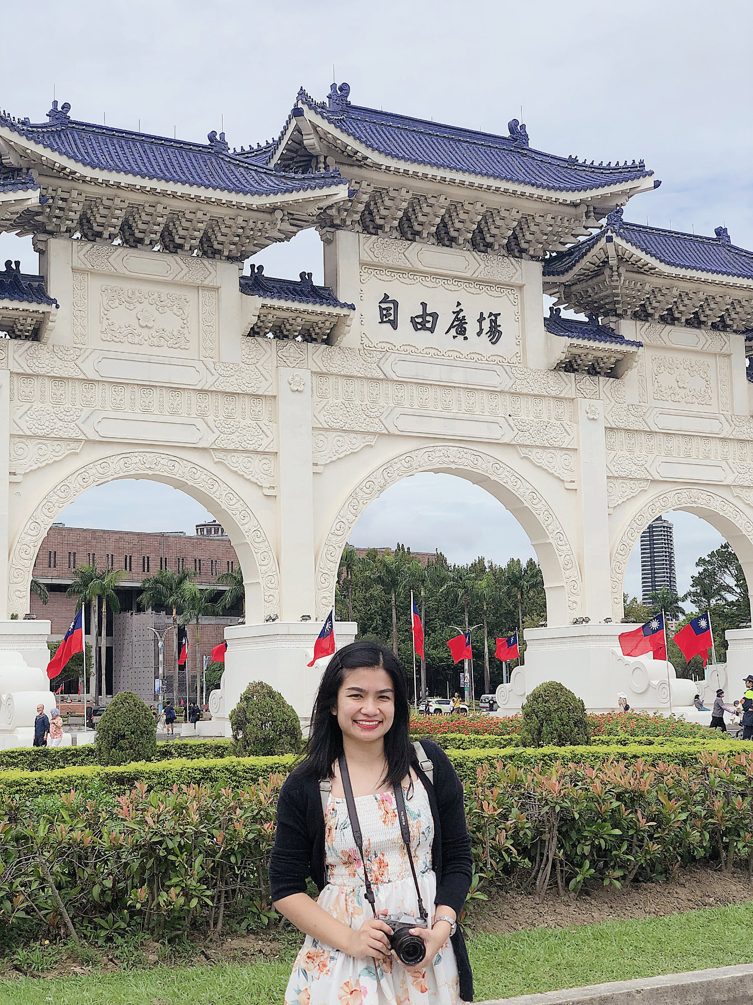 Young woman smiling, holding a camera, standing in front of a white traditional Chinese archway with Chinese characters and multiple Taiwanese flags, in a park with greenery and other visitors.