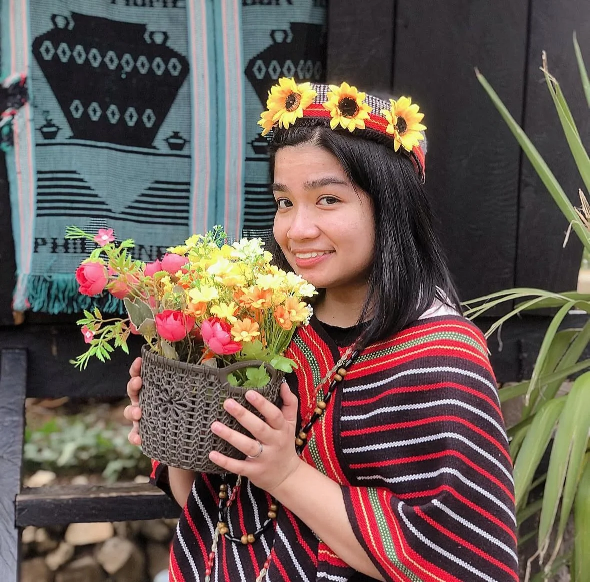 Young woman wearing traditional attire and sunflower crown, holding colorful flowers, smiling outdoors.