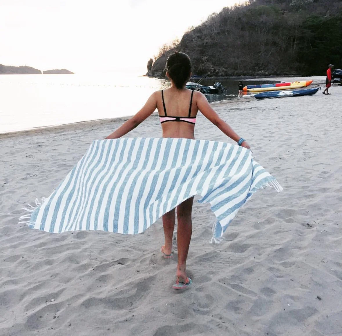 Woman walking on sandy beach holding a striped towel, with kayaks and a rocky headland in the background at sunset.