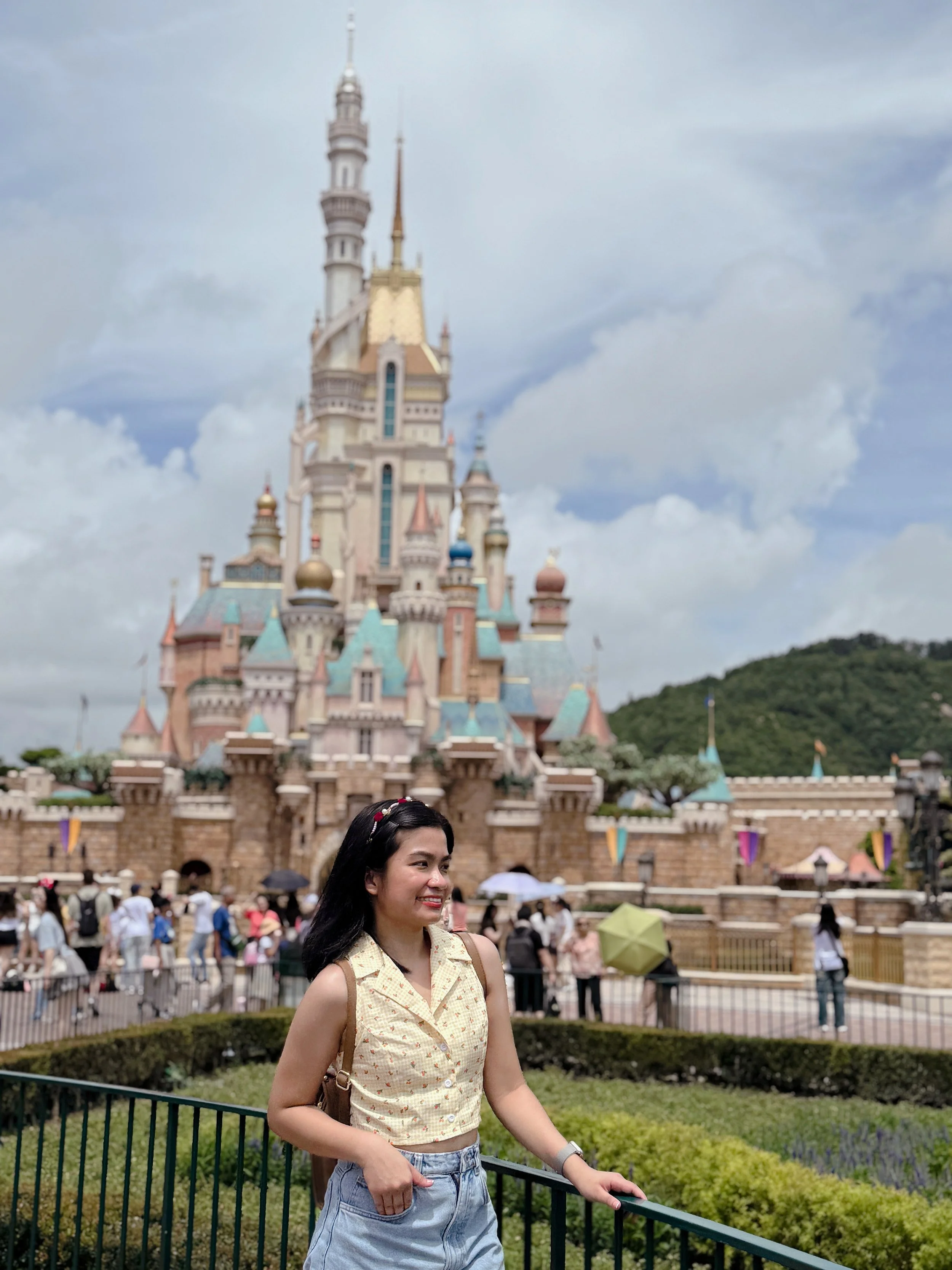 A woman smiling and standing by a fence in front of a castle at Disneyland.