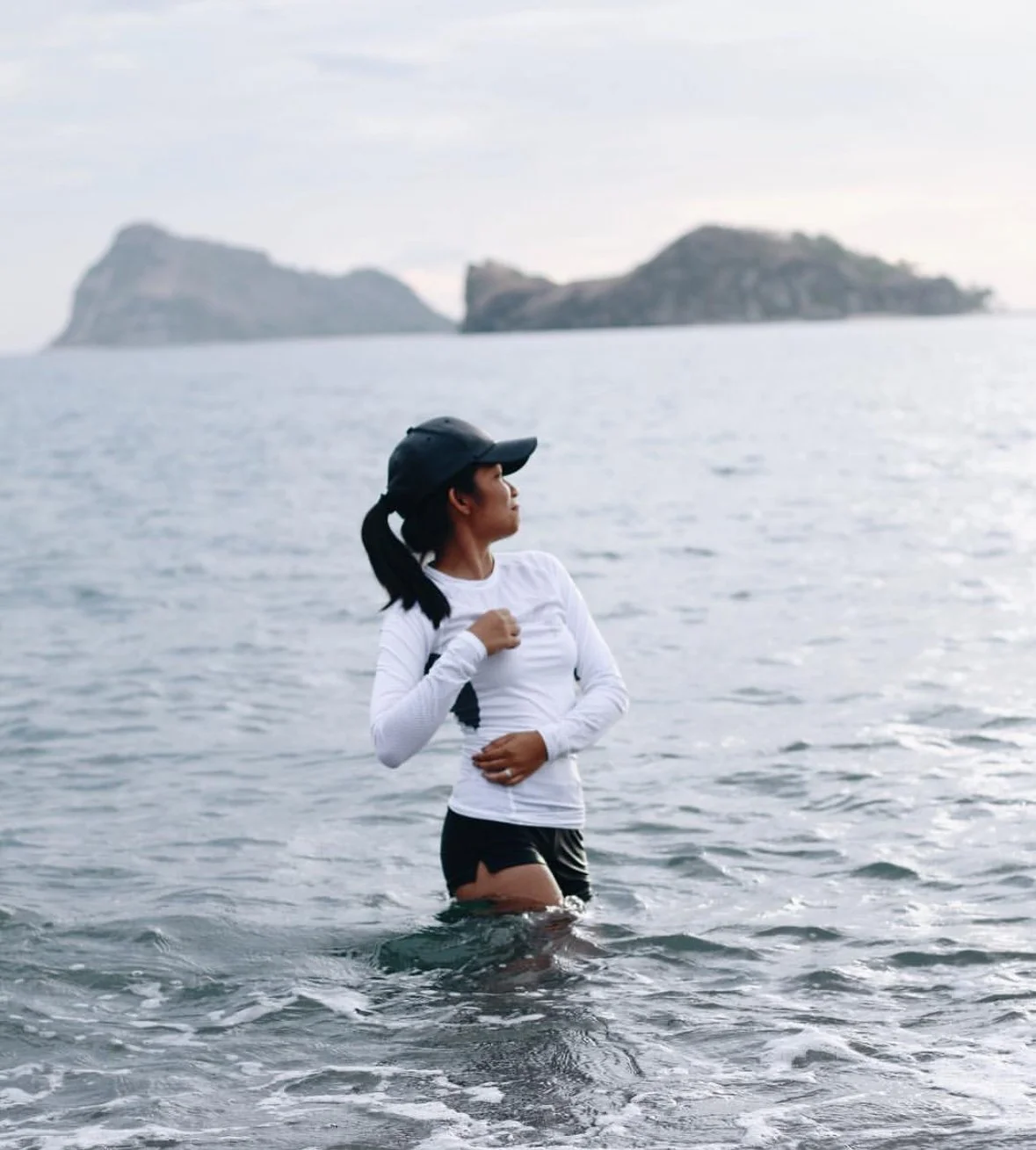 A woman standing in the ocean water near the shore with islands in the background, wearing a black cap, white long-sleeve shirt, and black shorts.