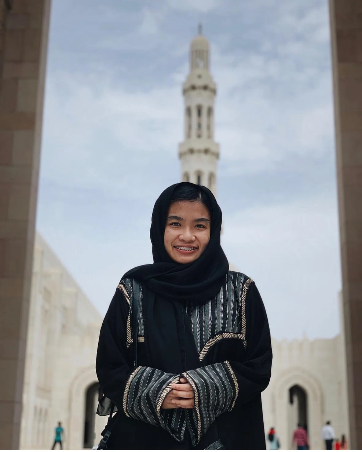 A young woman standing in front of a building with a clock tower, smiling at the camera. She is wearing a black headscarf and a black and patterned jacket. The background shows a partly cloudy sky and some people walking.