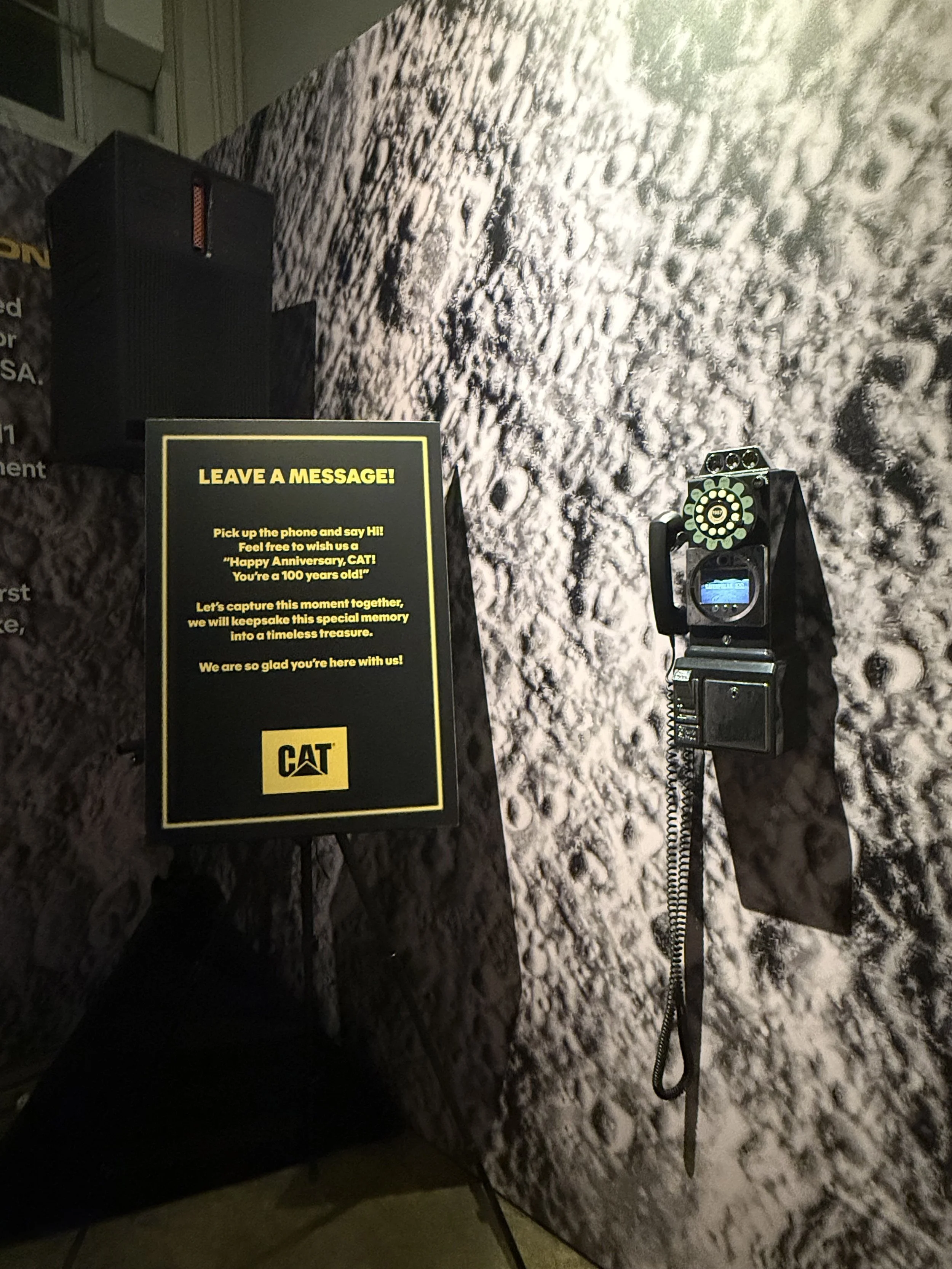 Photo of a moon landing exhibit featuring a vintage communication device mounted on a lunar surface backdrop, with a sign inviting visitors to leave a message.