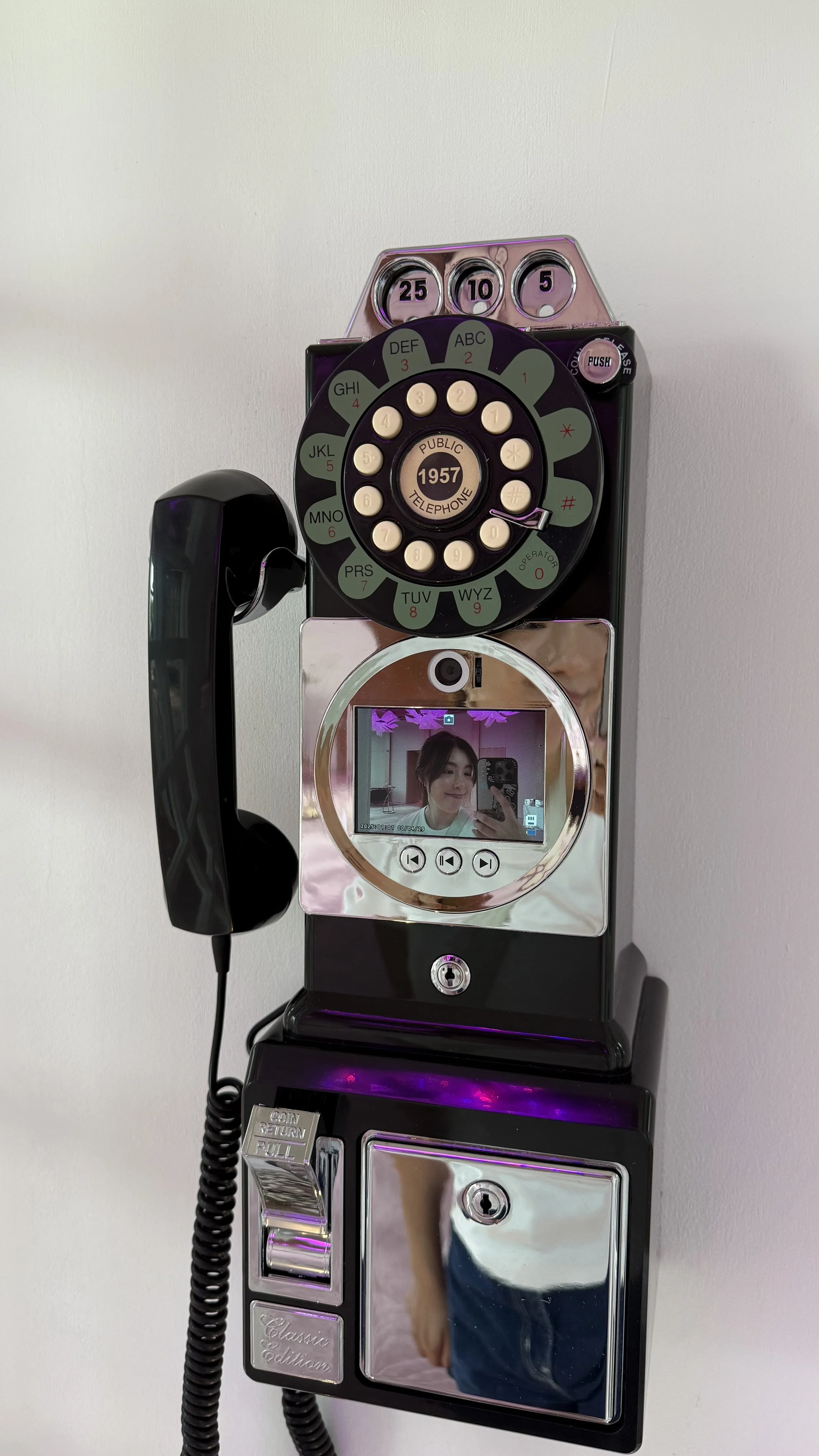 Vintage black payphone with a circular keypad, a rotary dial with a digital display, and a small camera lens. The phone is mounted on a wall with purple and silver accents, reflecting a person taking a selfie in the mirror.