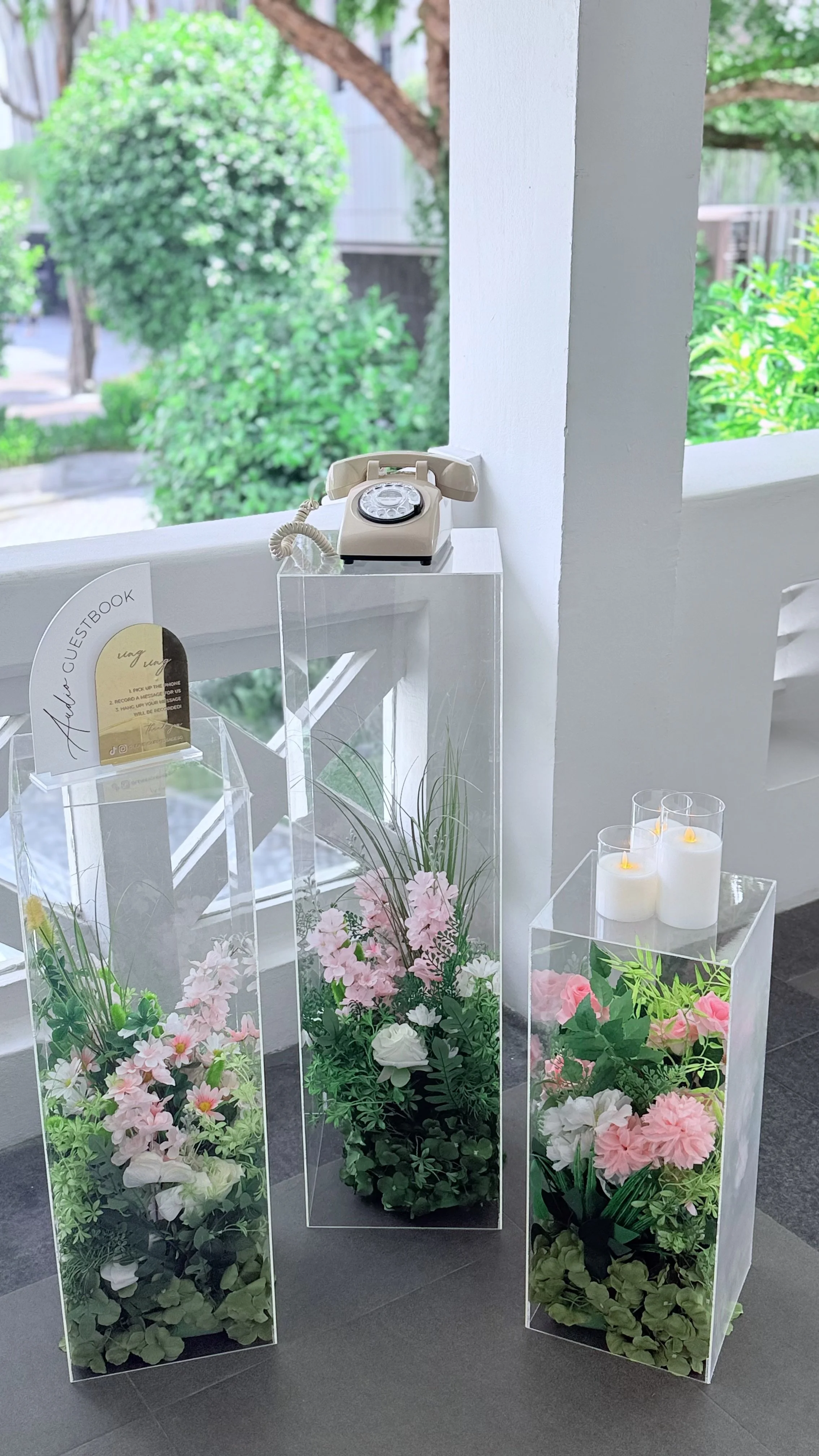 A vintage beige rotary telephone on a clear display stand, next to three pink bouquets in tall glass vases, and three white candles with yellow flames in glass holders, all set on a dark floor near a large window with an outdoor view of trees and greenery.