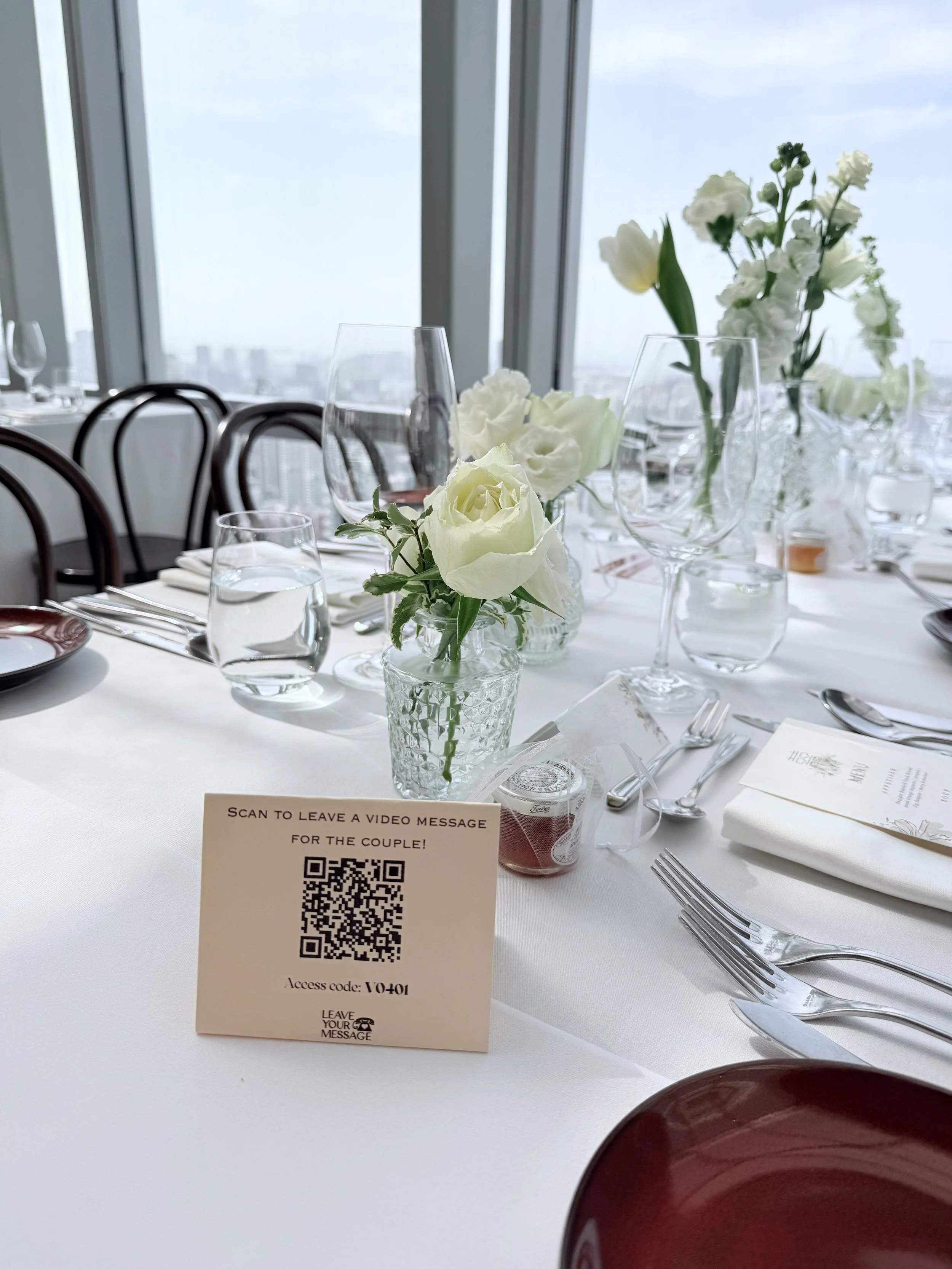Elegant banquet table set with white tablecloths, glassware, silverware, and floral centerpieces in a high-rise building with city views.