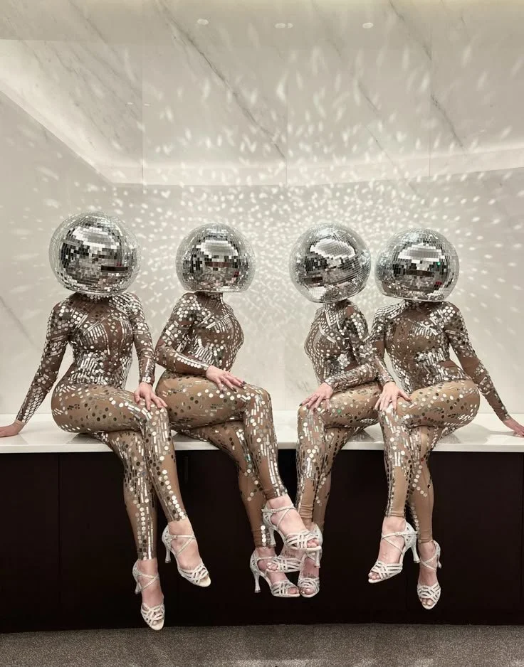 Four women in shiny, reflective bodysuits and high heels sit on a ledge with disco ball helmets, projecting light patterns on the wall behind them.