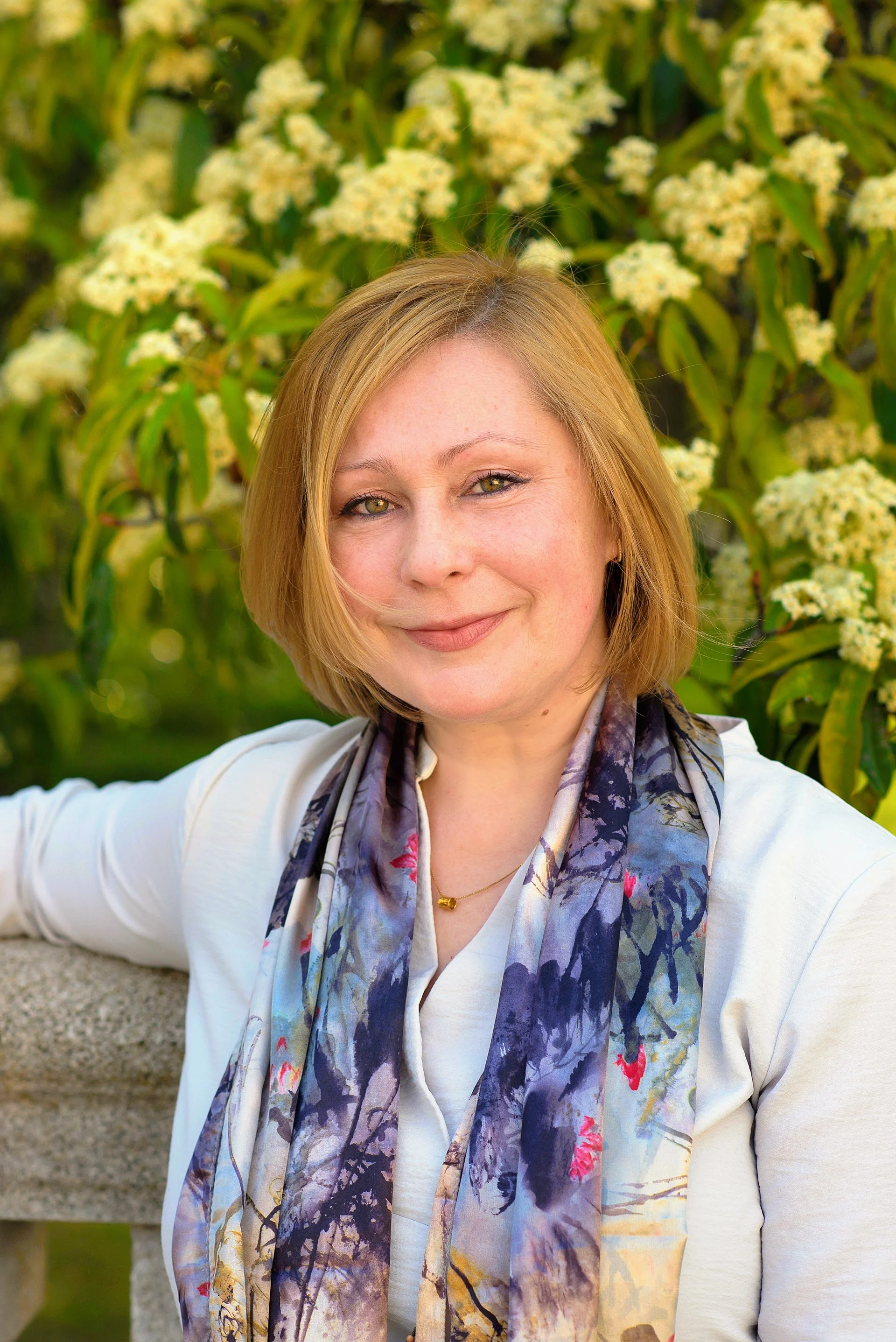 A woman with short blonde hair and green eyes, smiling outdoors with yellow flowers and green leaves in the background. She is wearing a white top, a multi-colored scarf, and a gold necklace.