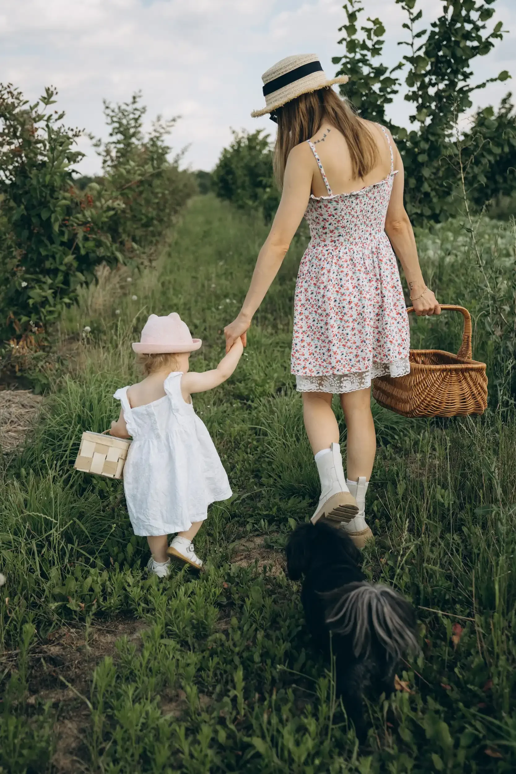 A woman and a young girl walking through a lush green orchard, holding hands. The woman is wearing a floral dress and a straw hat, and is carrying a wicker basket. The girl is dressed in a white dress and pink hat, holding a small basket. A dog walks ahead of them.