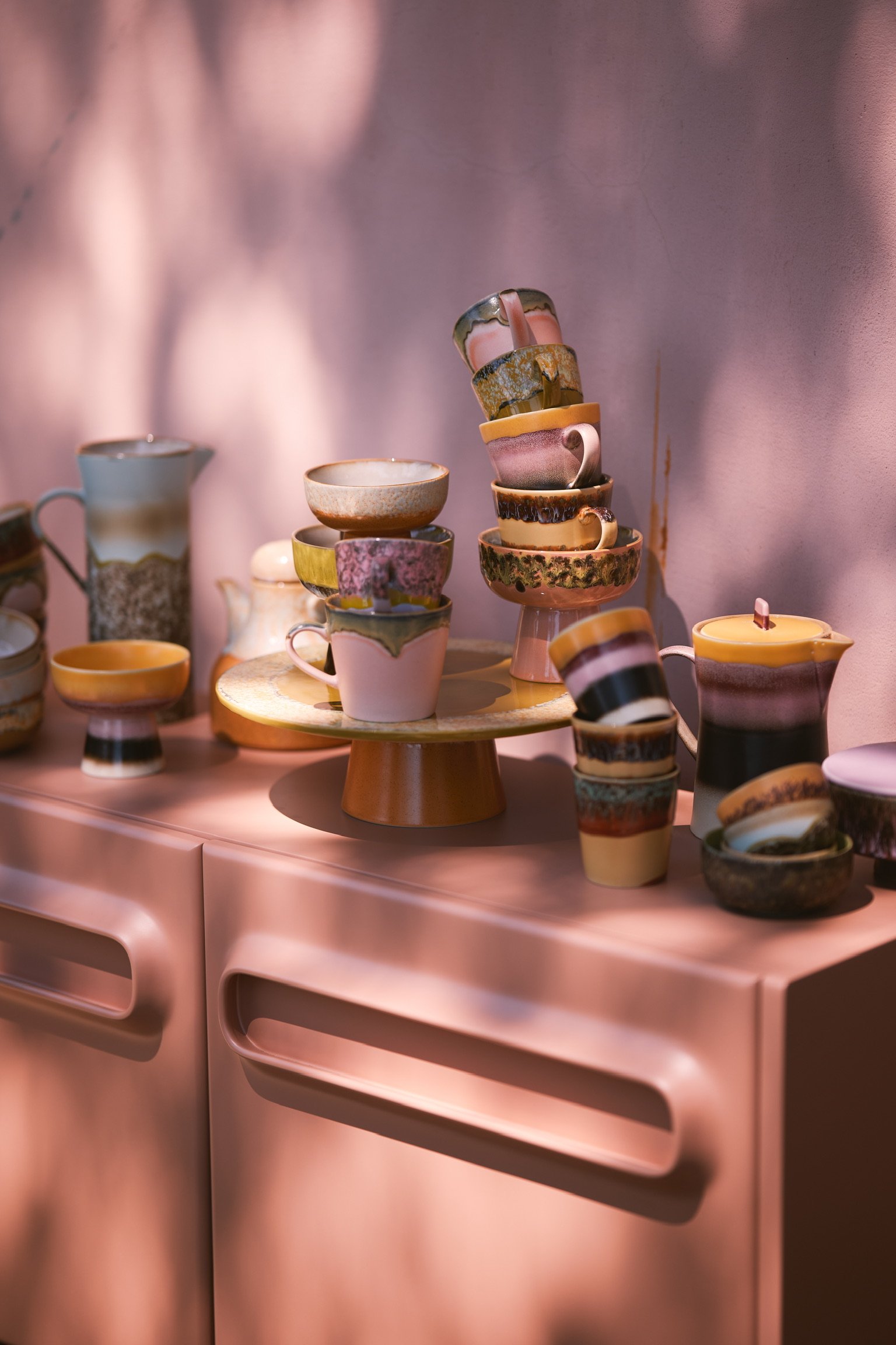 Eine Sammlung von bunten Keramik-Teetassen und -Schalen auf einem rosa Sideboard, einige gestapelt. Im Hintergrund eine rosa Wand mit Licht- und Schattenmustern.