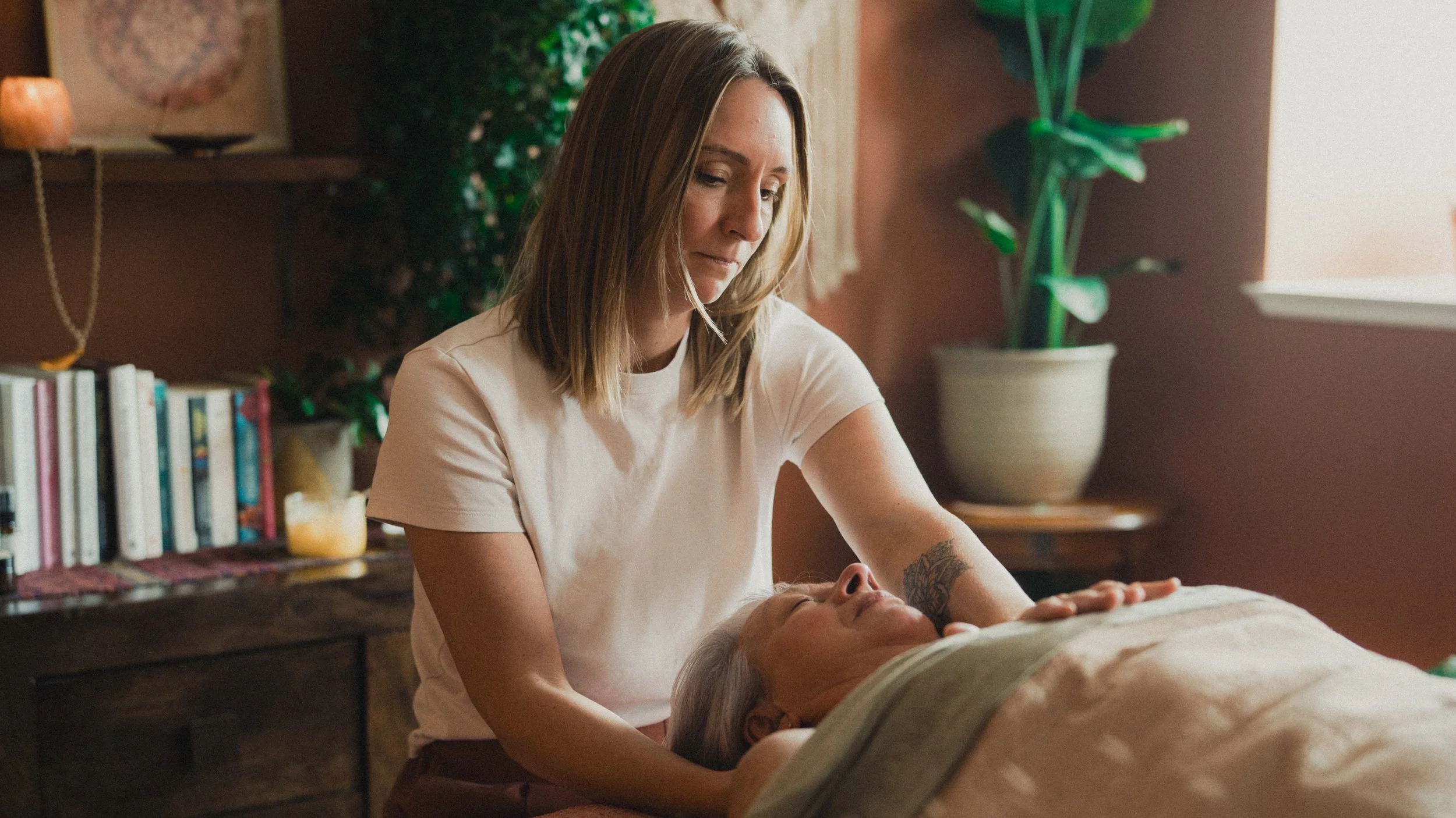 Woman comforting a person lying under a blanket in a warmly lit home with books and houseplants
