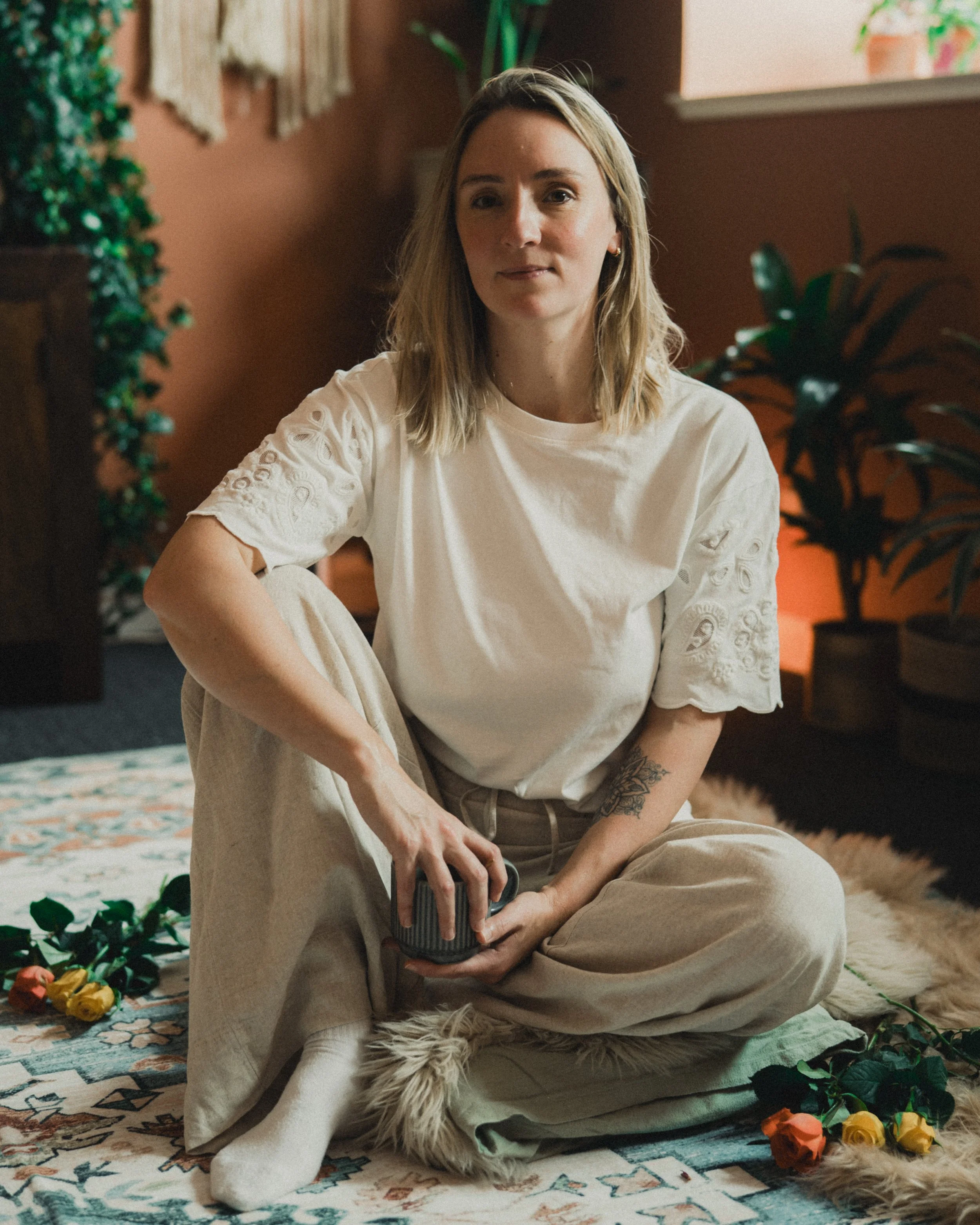 A woman with blonde hair sitting cross-legged on a patterned rug in a cozy room, holding a mug, surrounded by plants and colorful roses.