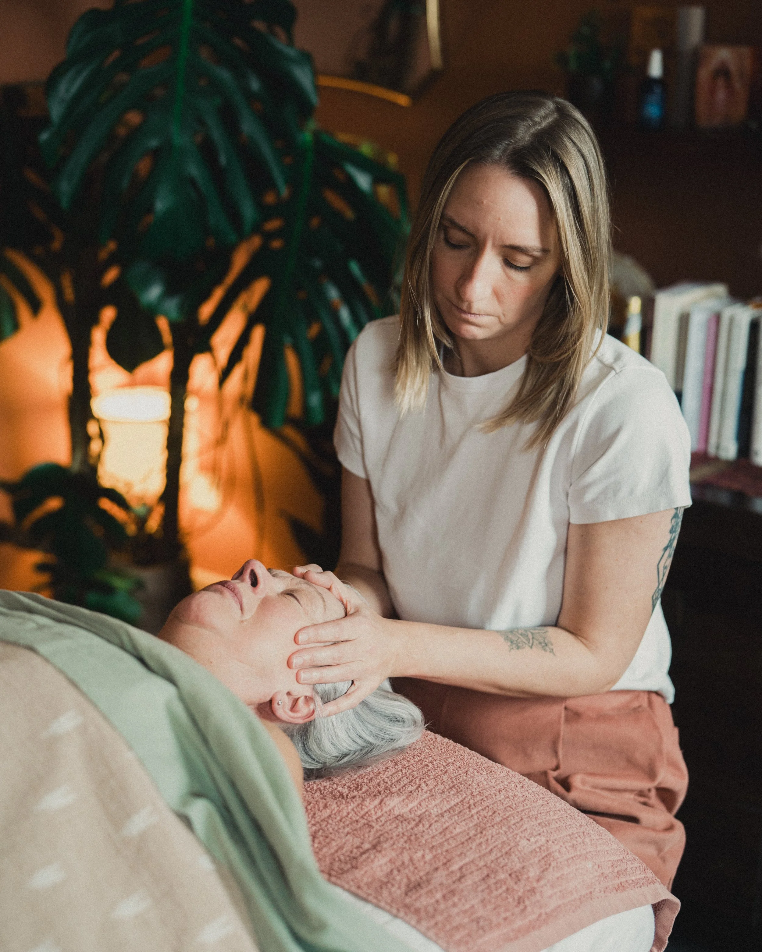 A woman giving a facial massage to an older woman lying on a massage table in a cozy, softly lit room.