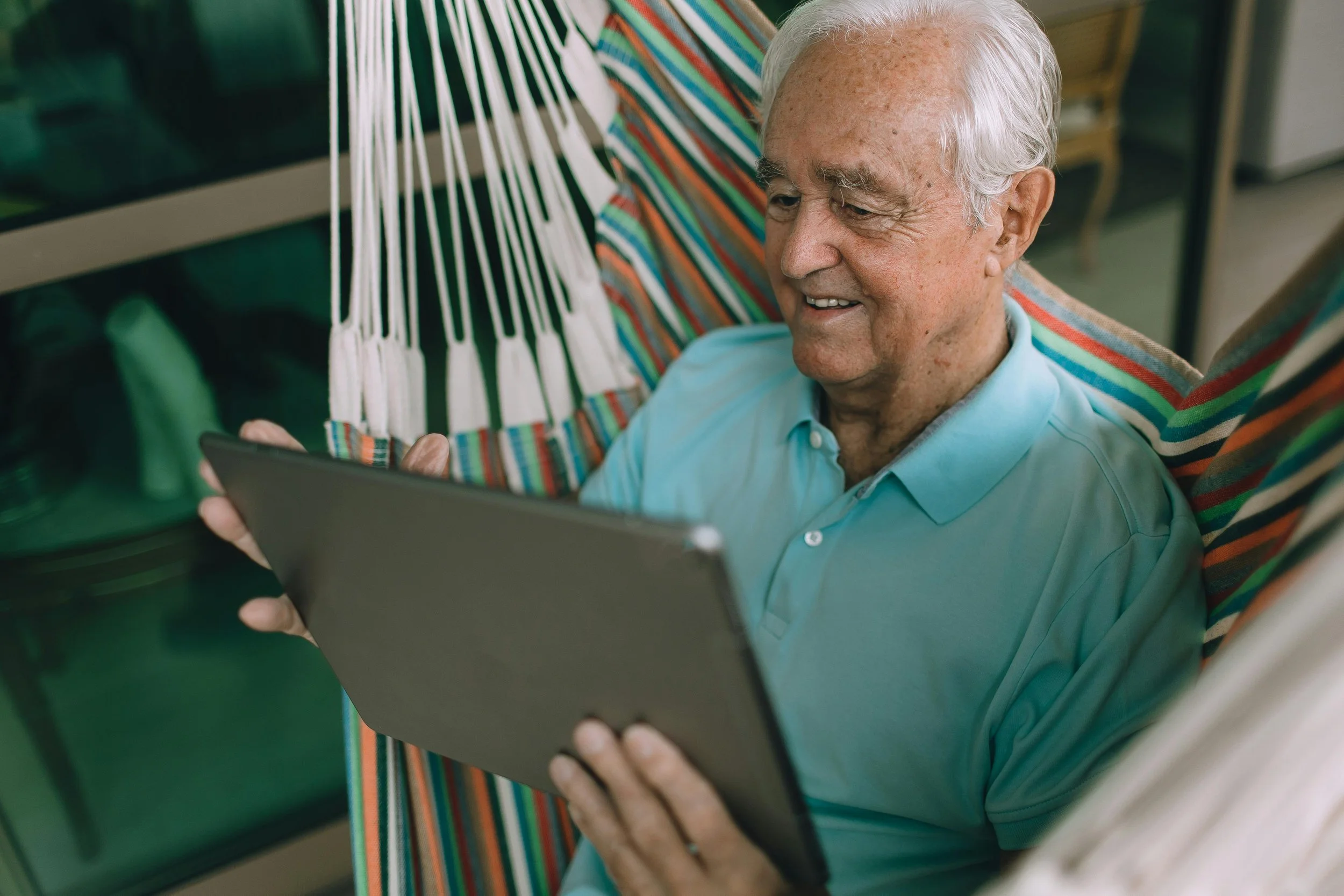 An elderly man sitting in a striped hammock, smiling and looking at a tablet device.