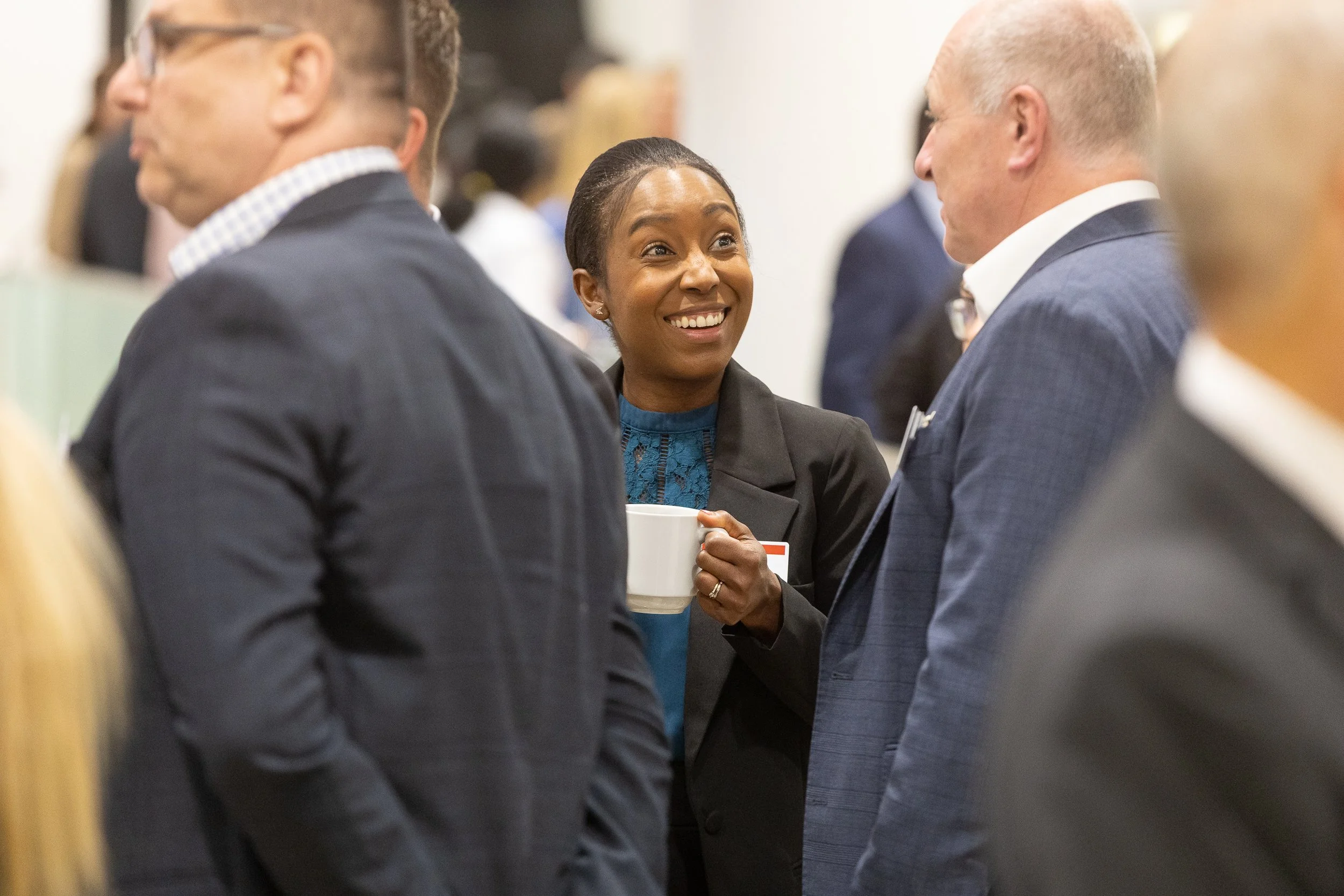A woman smiling and holding a mug while talking to two men in suits at a professional networking event.
