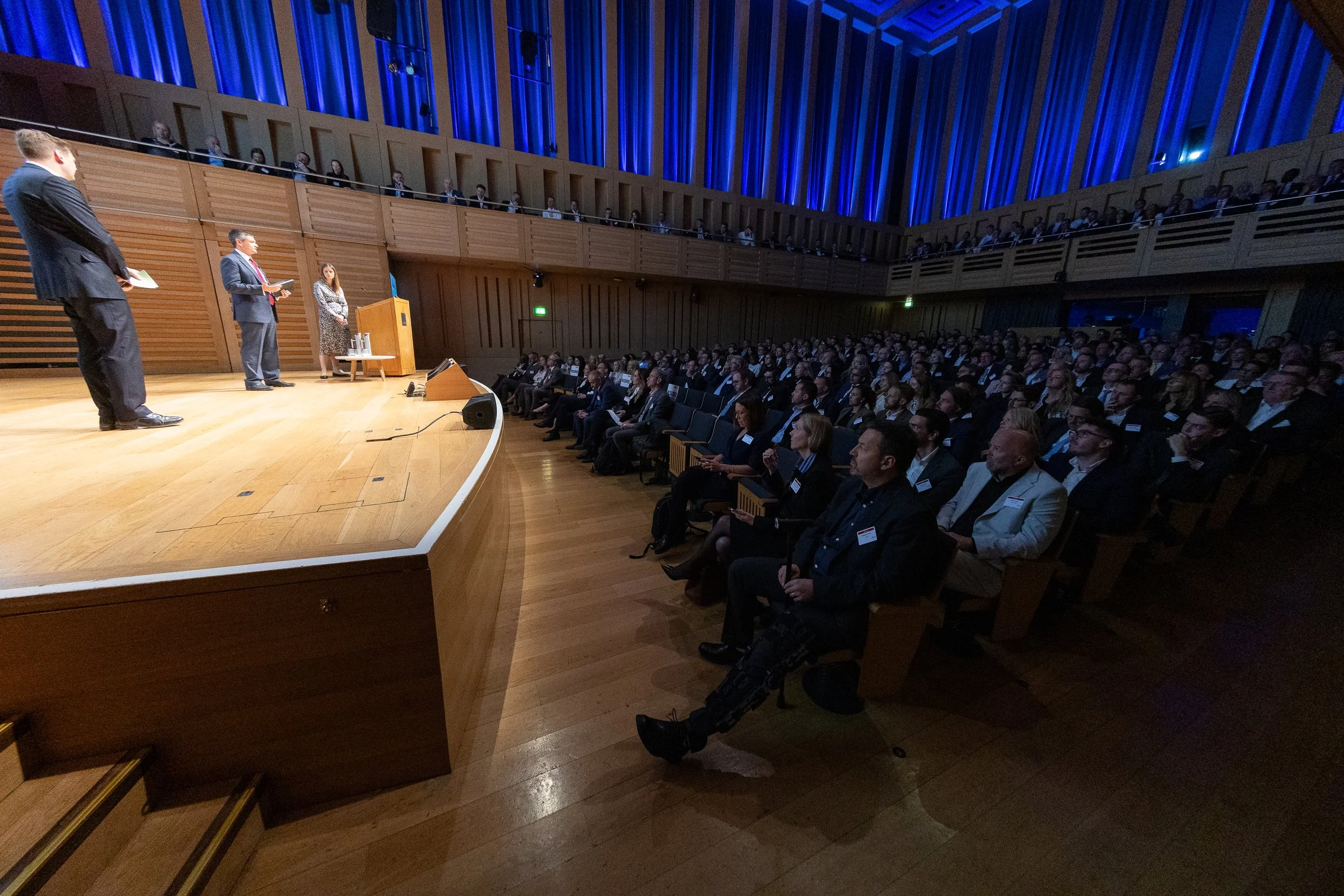 A large audience seated in an auditorium watching a panel of speakers presenting on stage, with a wooden floor, blue lighting, and multiple levels of seats.