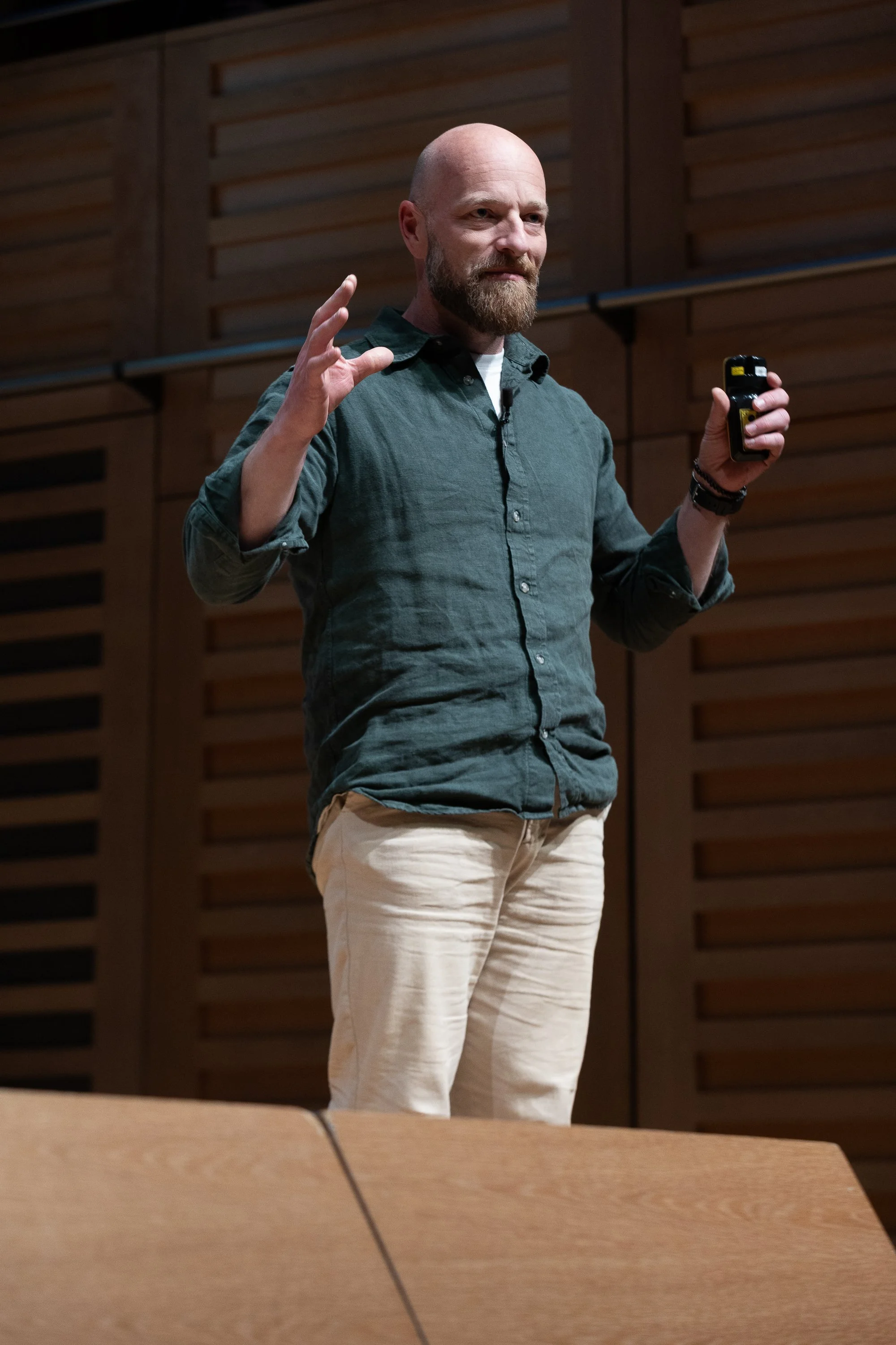 A man with a beard and bald head is giving a presentation in a room with wooden walls. He is wearing a dark green button-up shirt with the sleeves rolled up and light-colored pants, holding a small black device in his right hand.