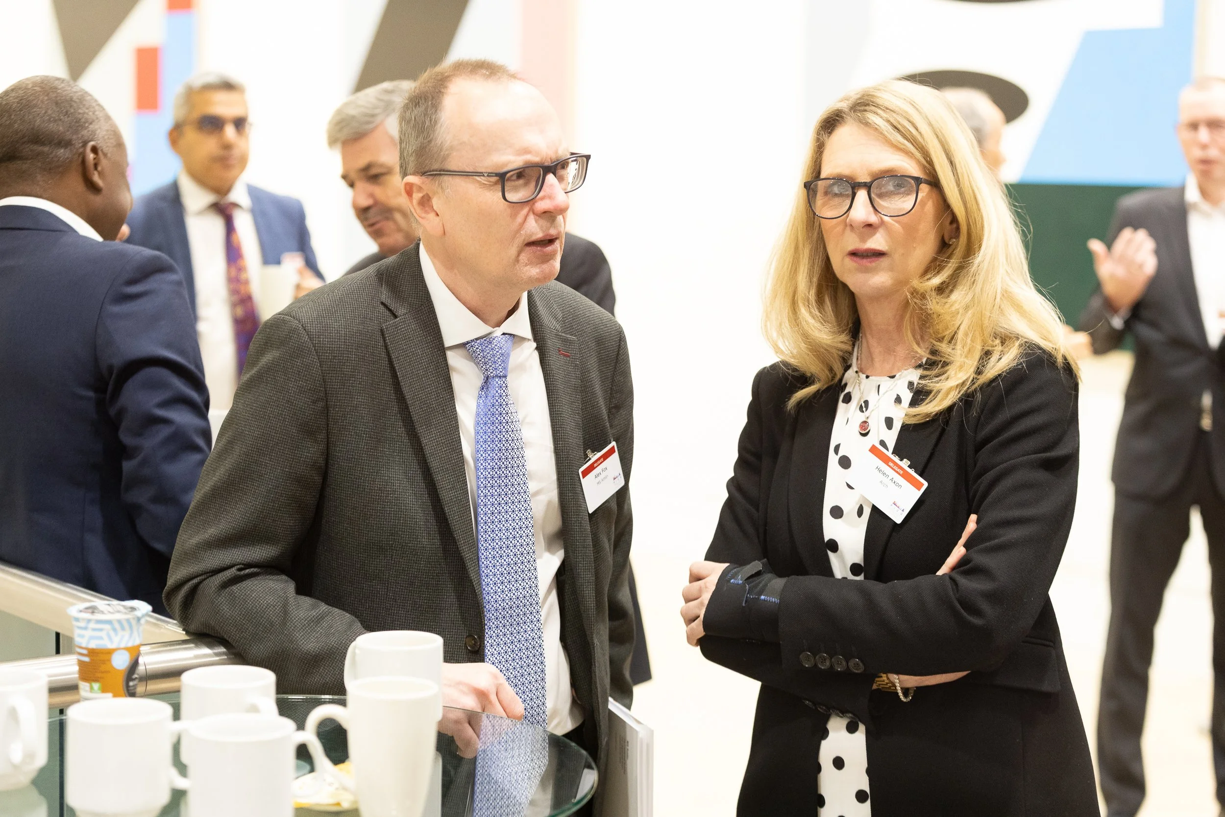 A group of business professionals having a conversation at a networking event, with a woman in a black blazer and polka dot blouse and a man in a gray suit and glasses at the center.