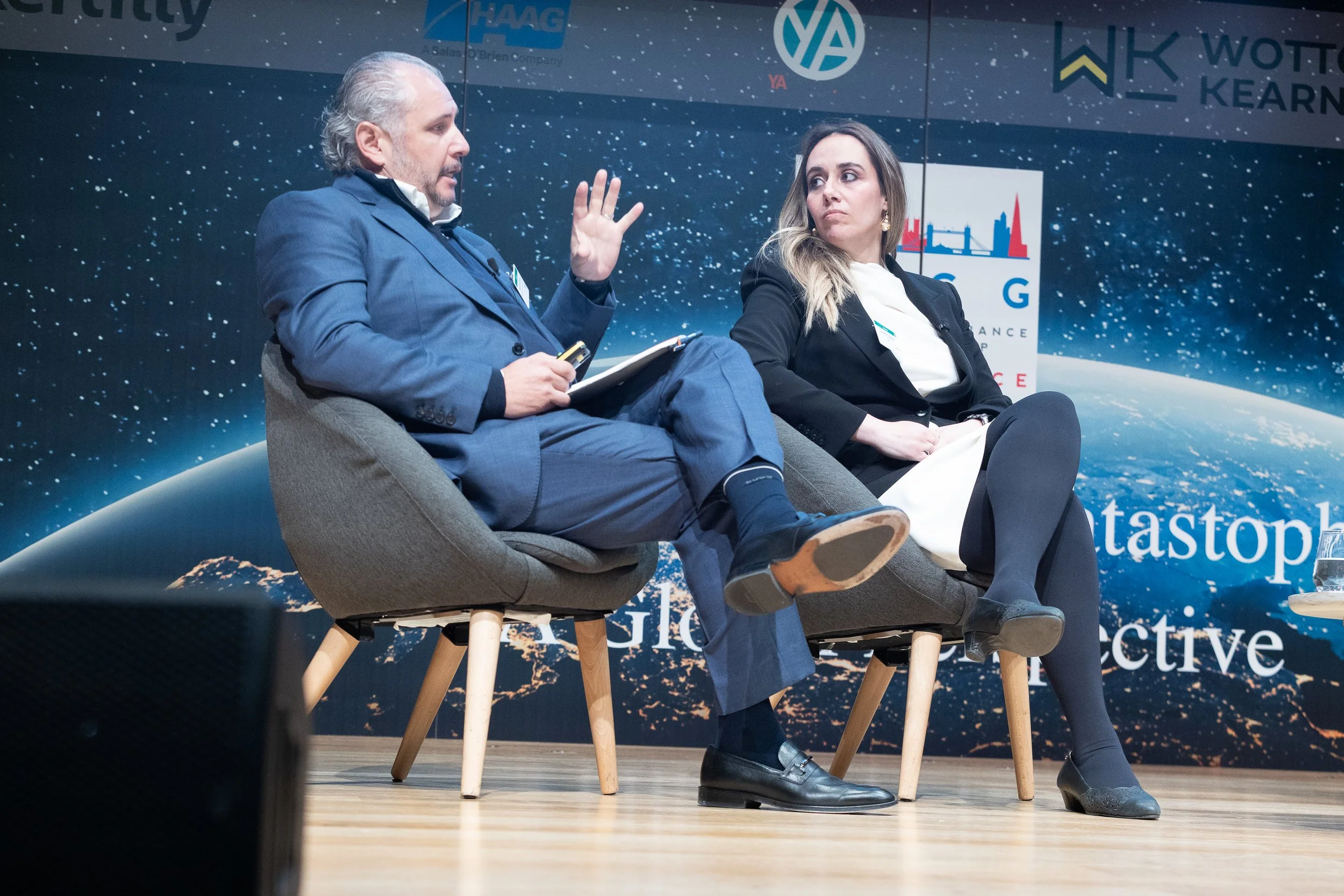 A man and woman seated on a stage, engaged in a conversation during a conference or panel discussion. The background features space-themed graphics and event branding.