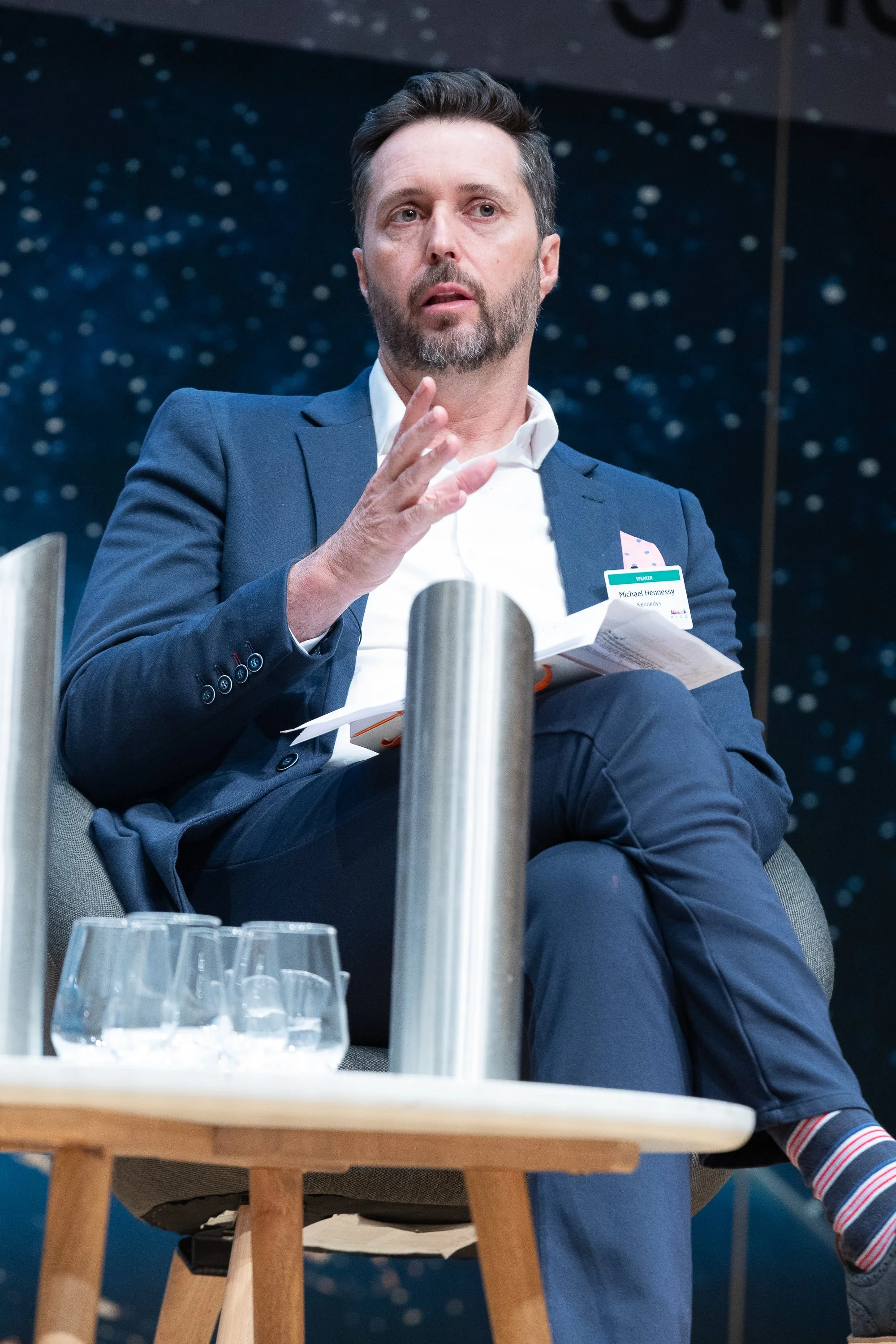 A man in a suit sitting on a chair, speaking or gesturing during a panel or conference. He has a beard, short hair, and is holding papers. There is a table with glasses and a backdrop with a starry sky pattern.