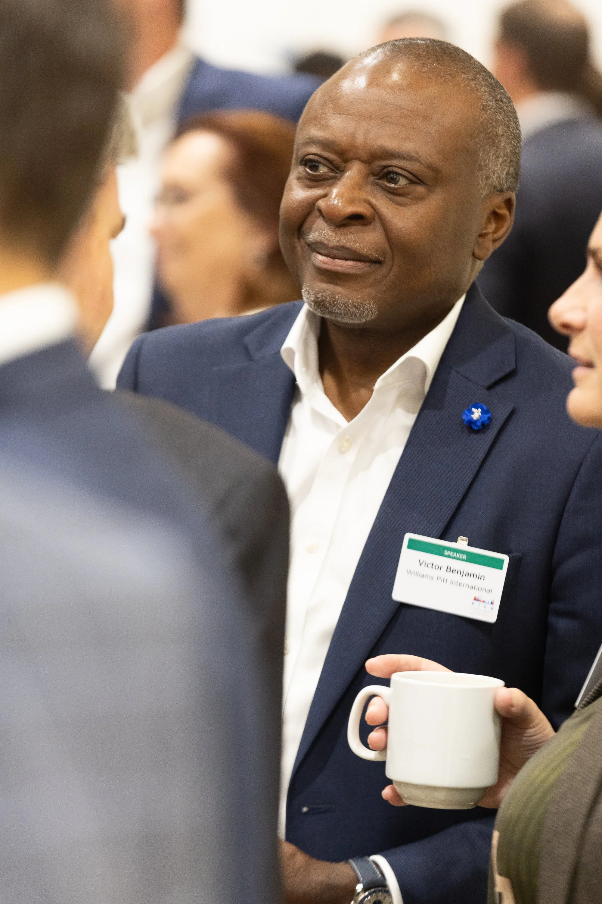 A man in a navy suit and white shirt, with a name badge that reads Victor Benjamin, is conversing with a group at a professional event, holding a white mug in his hand.