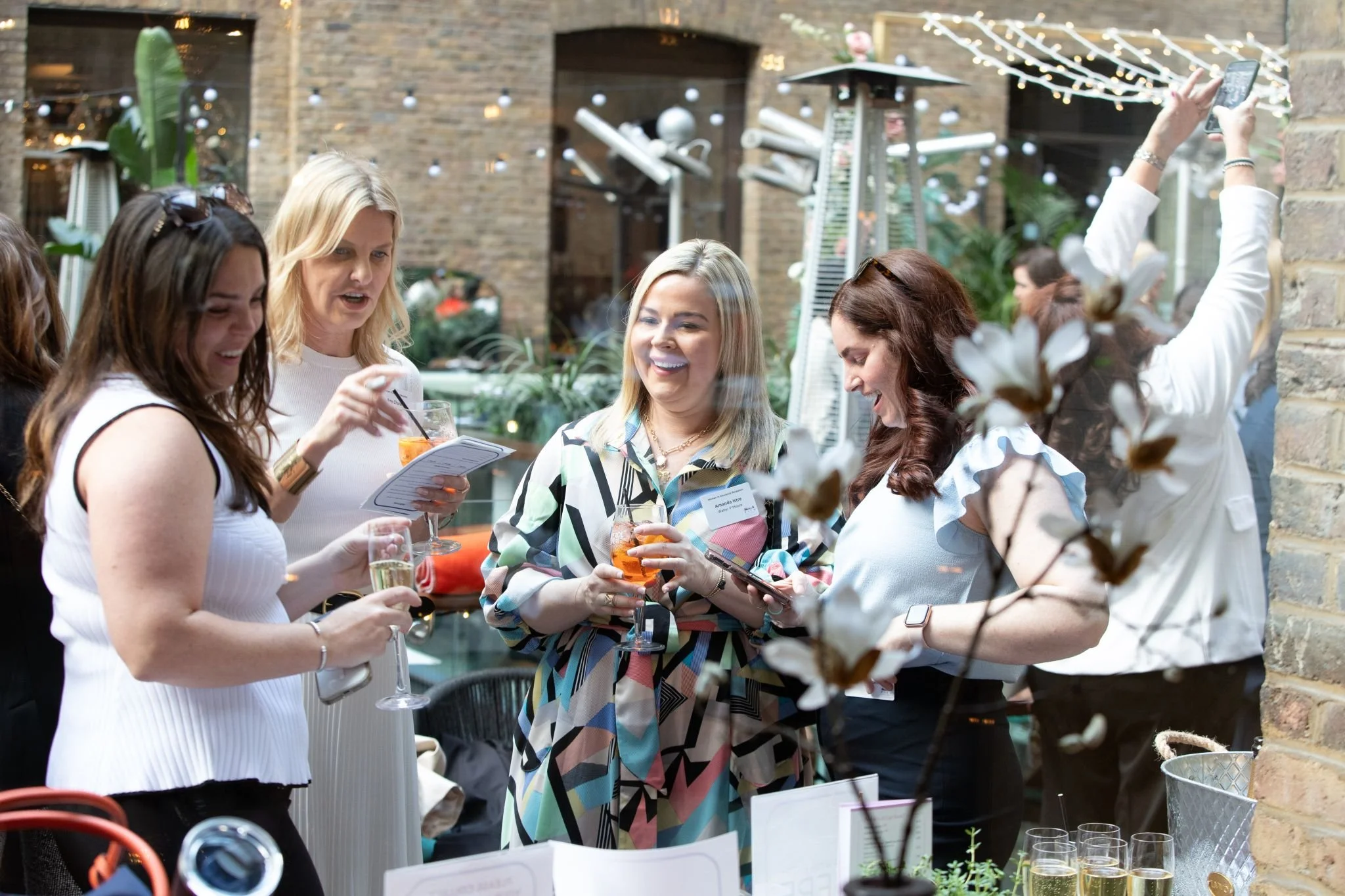 Group of women socializing at a party, holding drinks, smiling, and looking at their phones in an outdoor setting with string lights and plants.