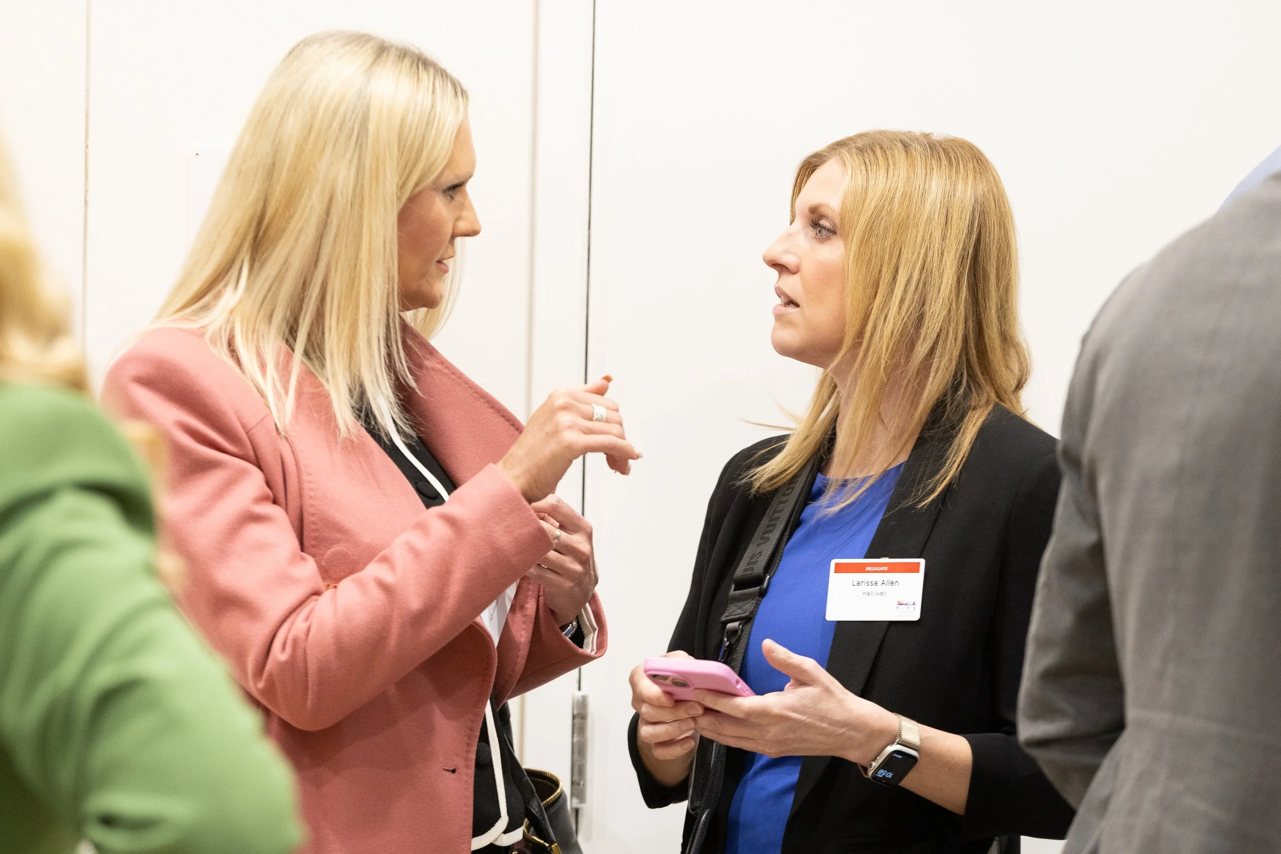 Two women in conversation at a professional event, one gesturing with her hand and the other holding a pink phone, one woman has a name tag reading 'Larissa Allen'.
