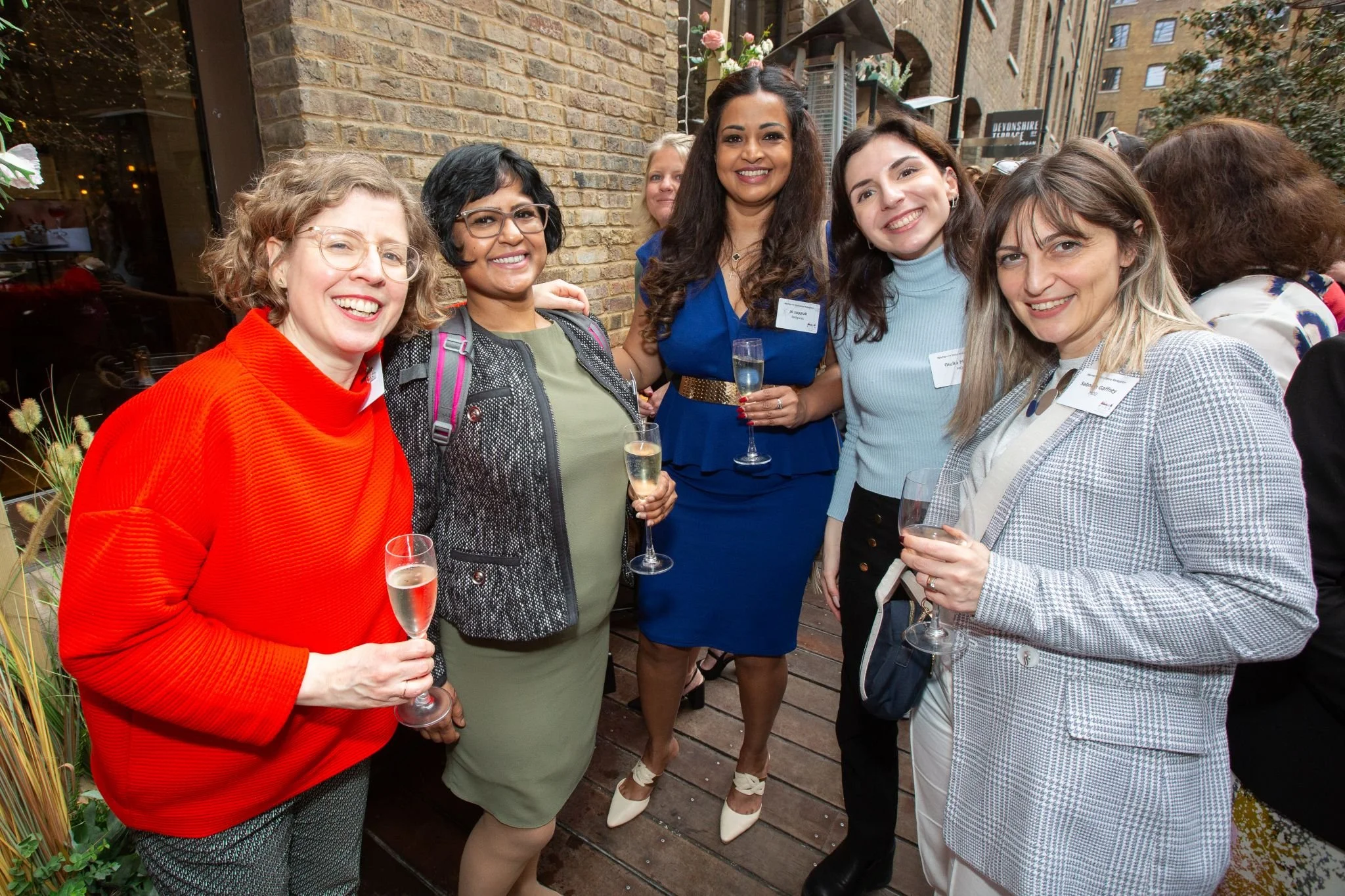 Group of women at a social event holding champagne glasses, standing on a wooden deck outside a brick building.