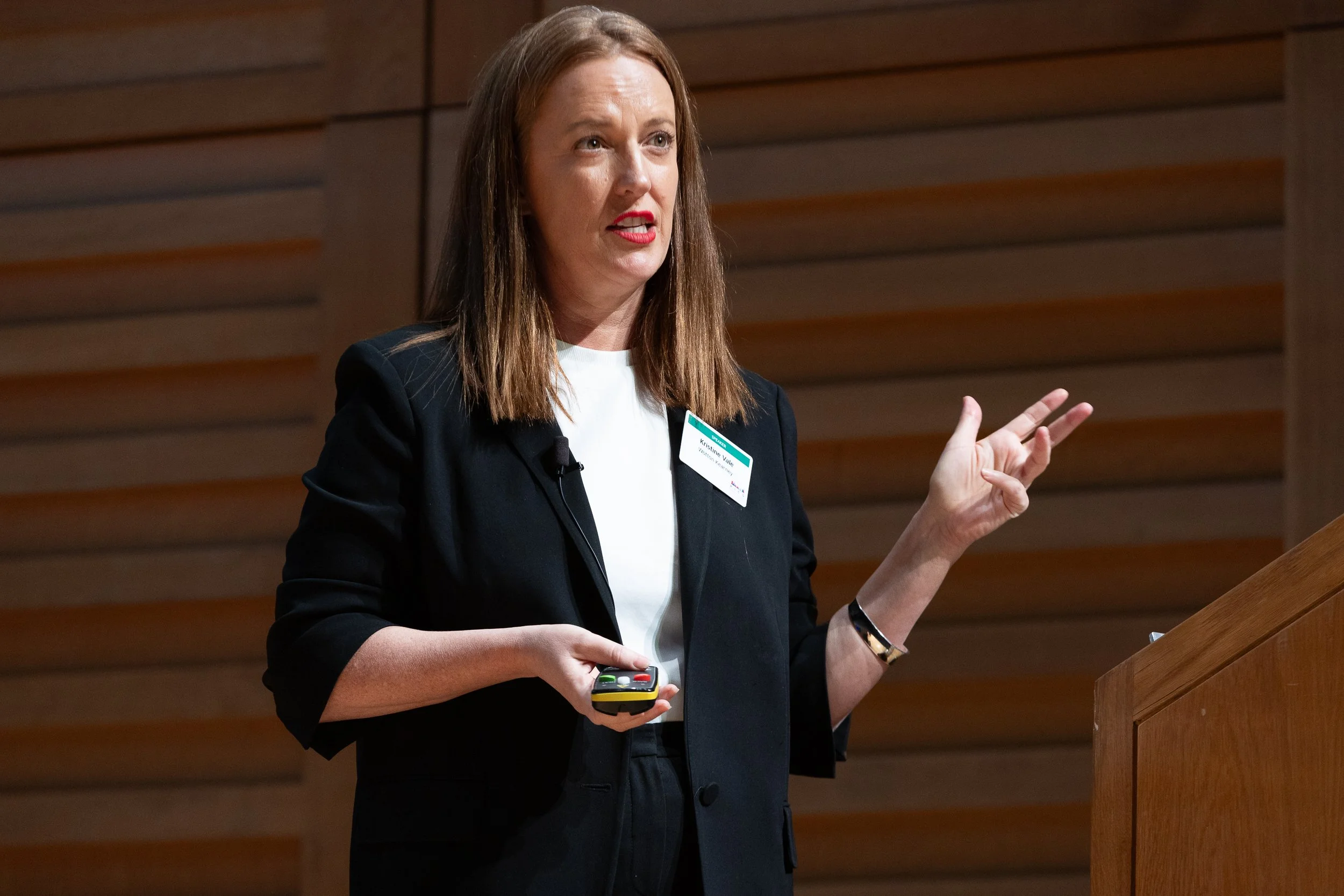 A woman with shoulder-length brown hair wearing a black blazer and white top, standing behind a podium, giving a presentation, holding a remote control in her left hand, gesturing with her right hand, with a wood-paneled background.
