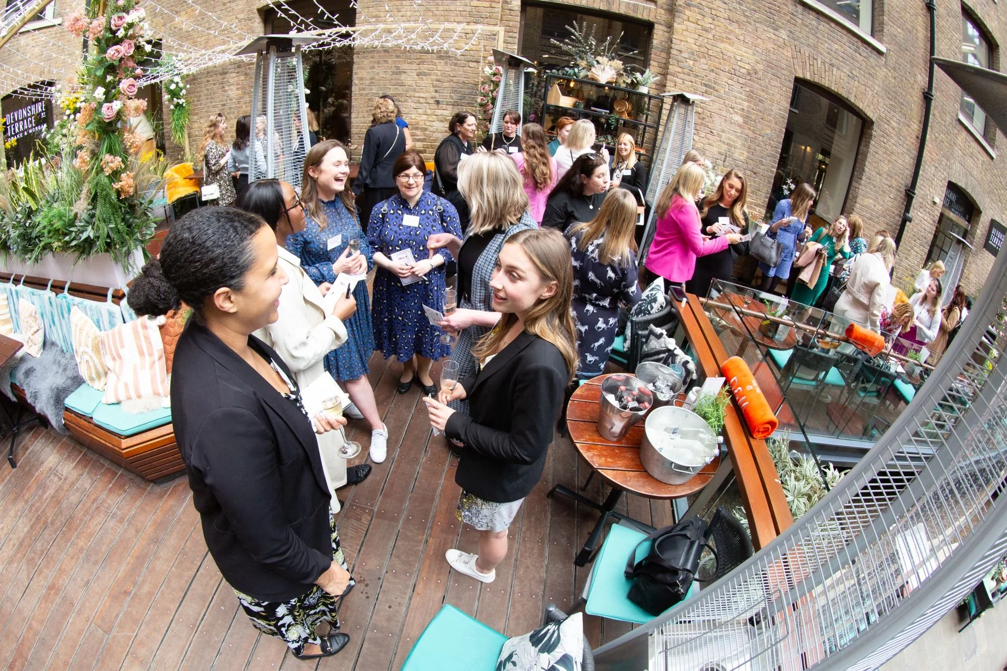 Women socializing at an outdoor venue with plants, drinks, and decorations, on a wooden deck.