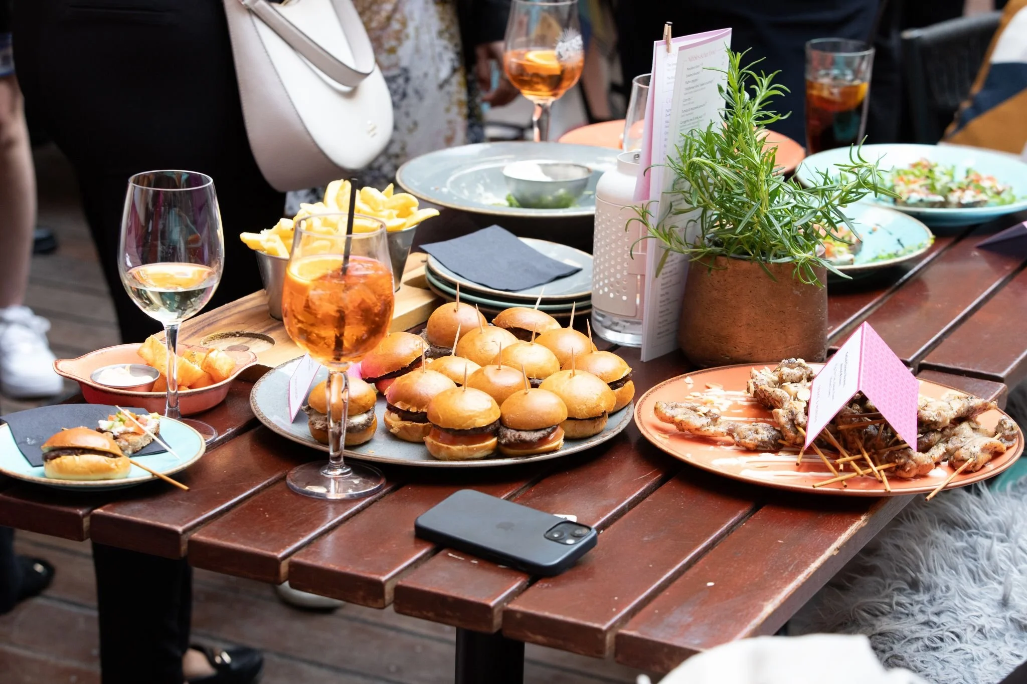 A wooden outdoor table with various appetizers and drinks, including sliders, chicken wings, small sandwiches on skewers, and glasses of wine and rosé, with a potted plant and menus.