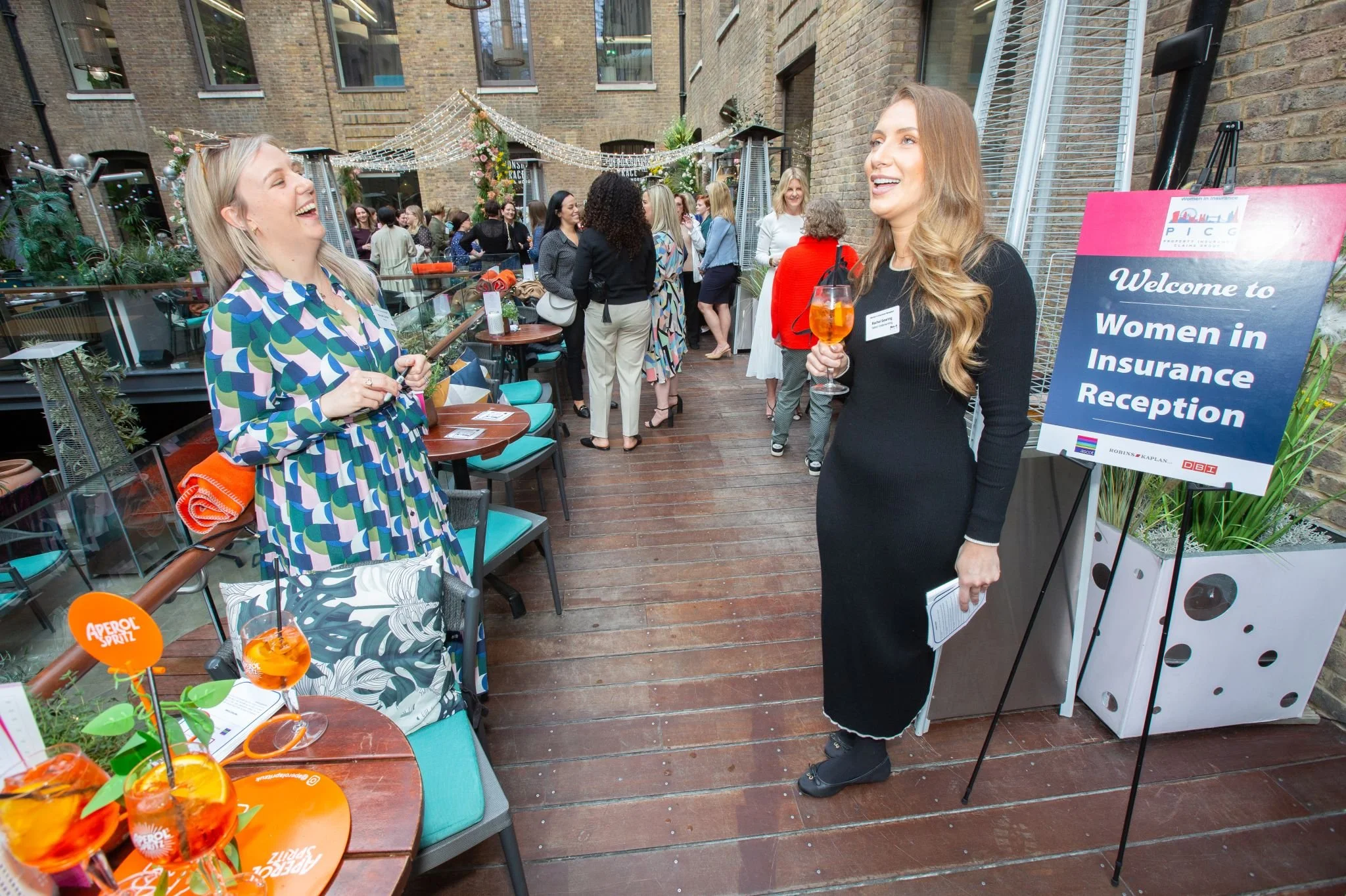 Two women are standing and conversing at an outdoor women in insurance reception event, holding drinks, with other attendees in the background on a decorated patio.