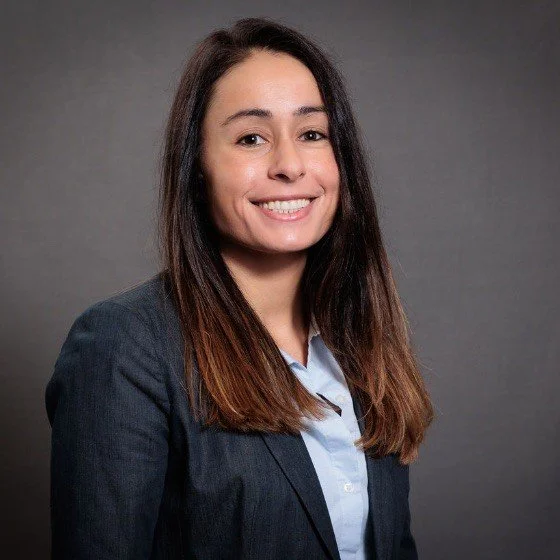 Smiling woman with shoulder-length black hair wearing a blue top in a greenhouse with green plants in the background.