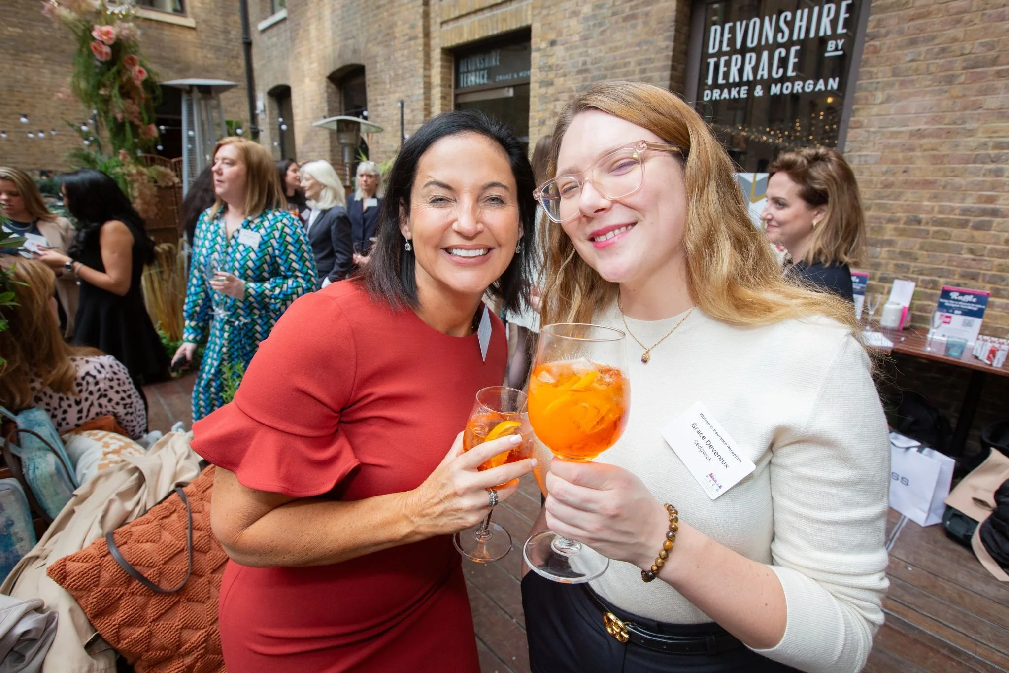Two smiling women holding glasses of orange drink at a social event, standing close together with other people in the background, in an indoor venue with brick walls and a sign that reads 'Devonshire Terrace by Drake & Morgan'.