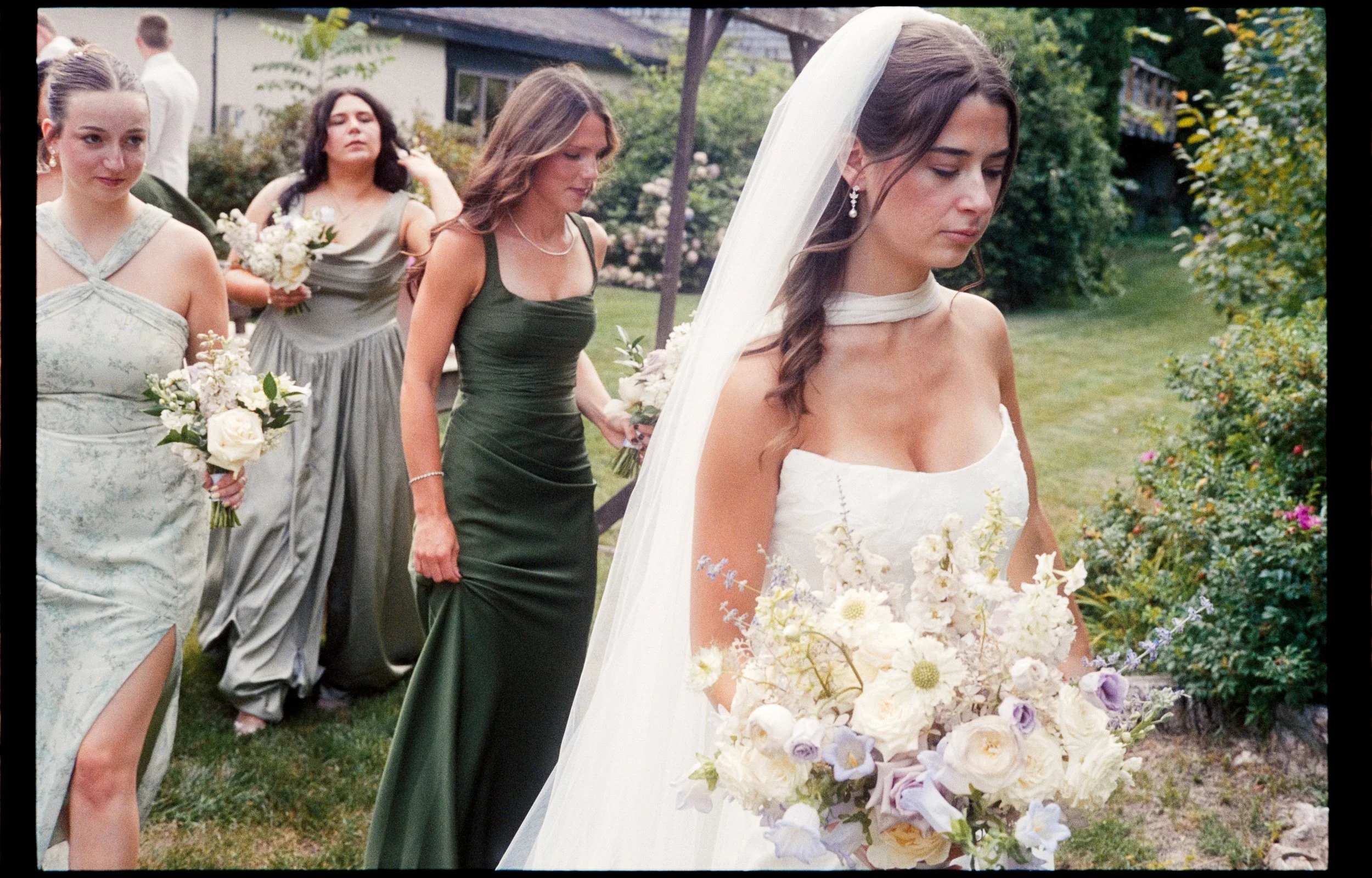 A bride in a white wedding dress and veil holding a large bouquet of white and pastel flowers, walking outdoors with bridesmaids and guests at a garden wedding.