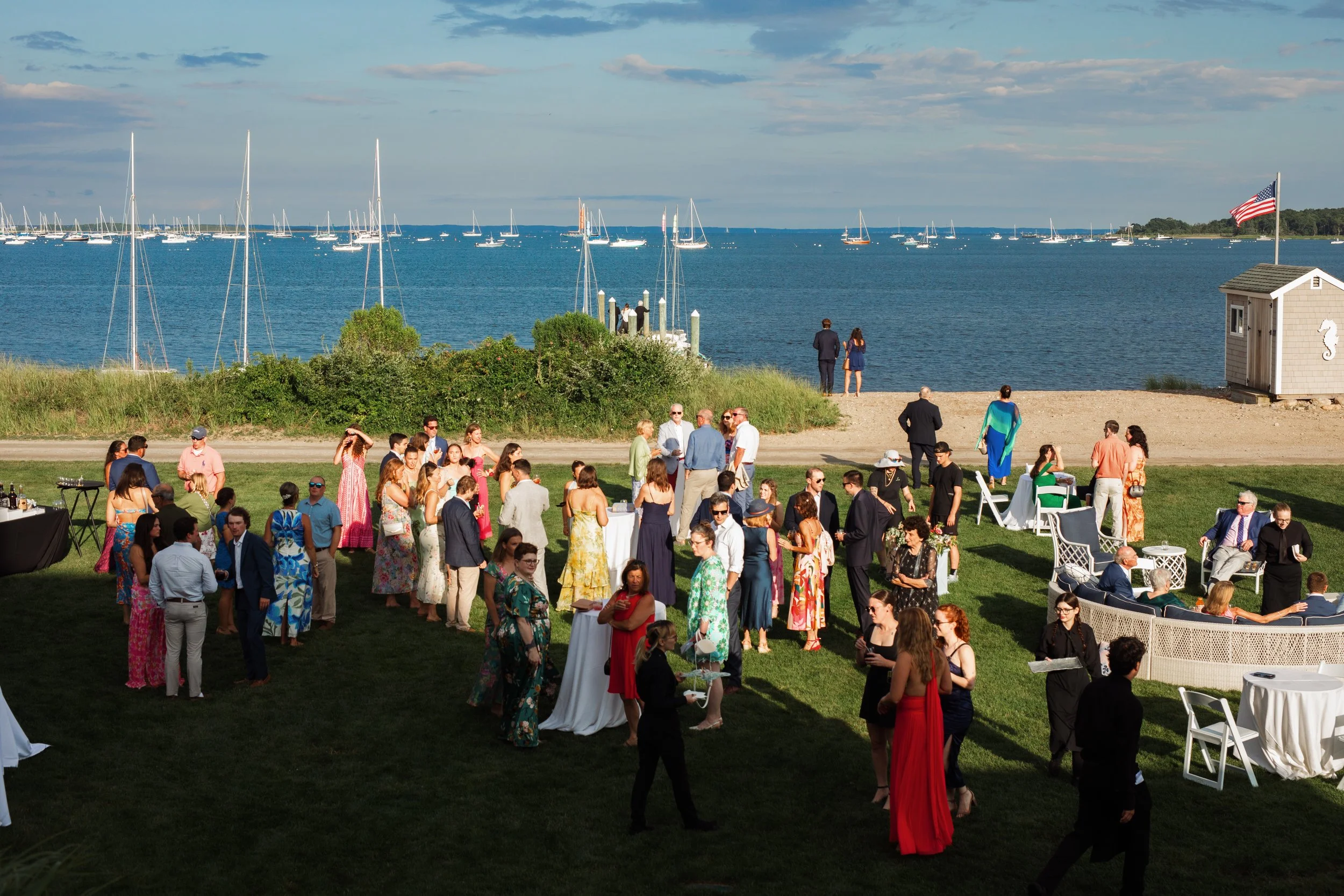 Guests in formal attire gather together during cocktail hour over looking the harbor filled with sail boats.