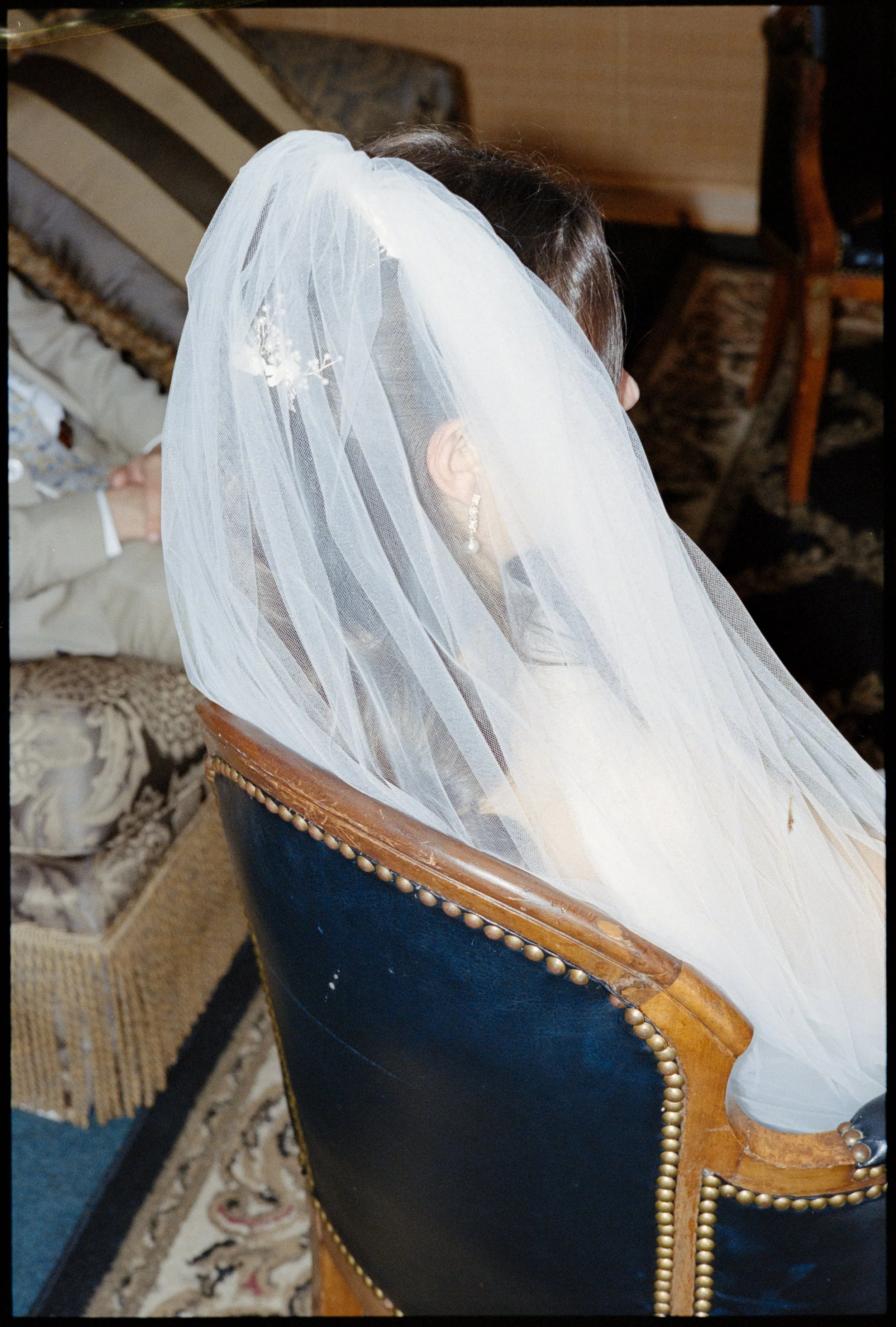 A bride sits in an antique leather and wood chair and her veil is tucked in behind her.