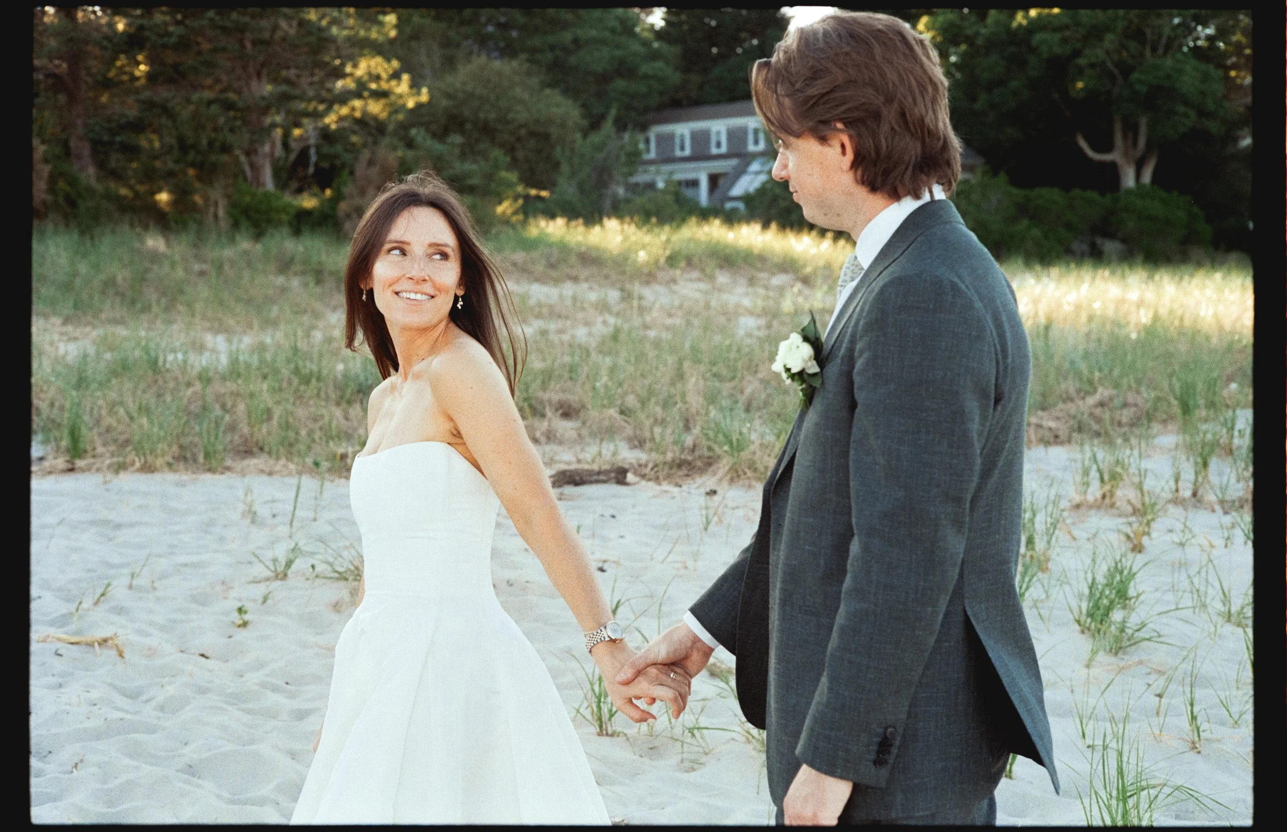 A smiling woman in a white dress holding hands with a man in a gray suit and tie, on a sandy beach with green grass and trees in the background.