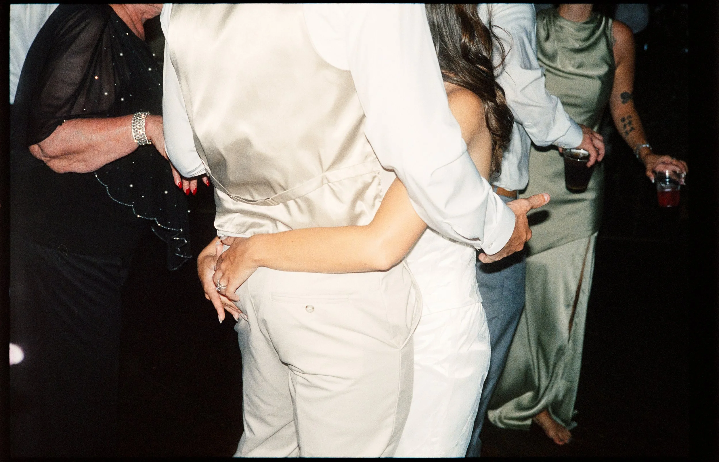 A couple embraces as guests dance and celebrate around the couple in satin and bedazzled clothing